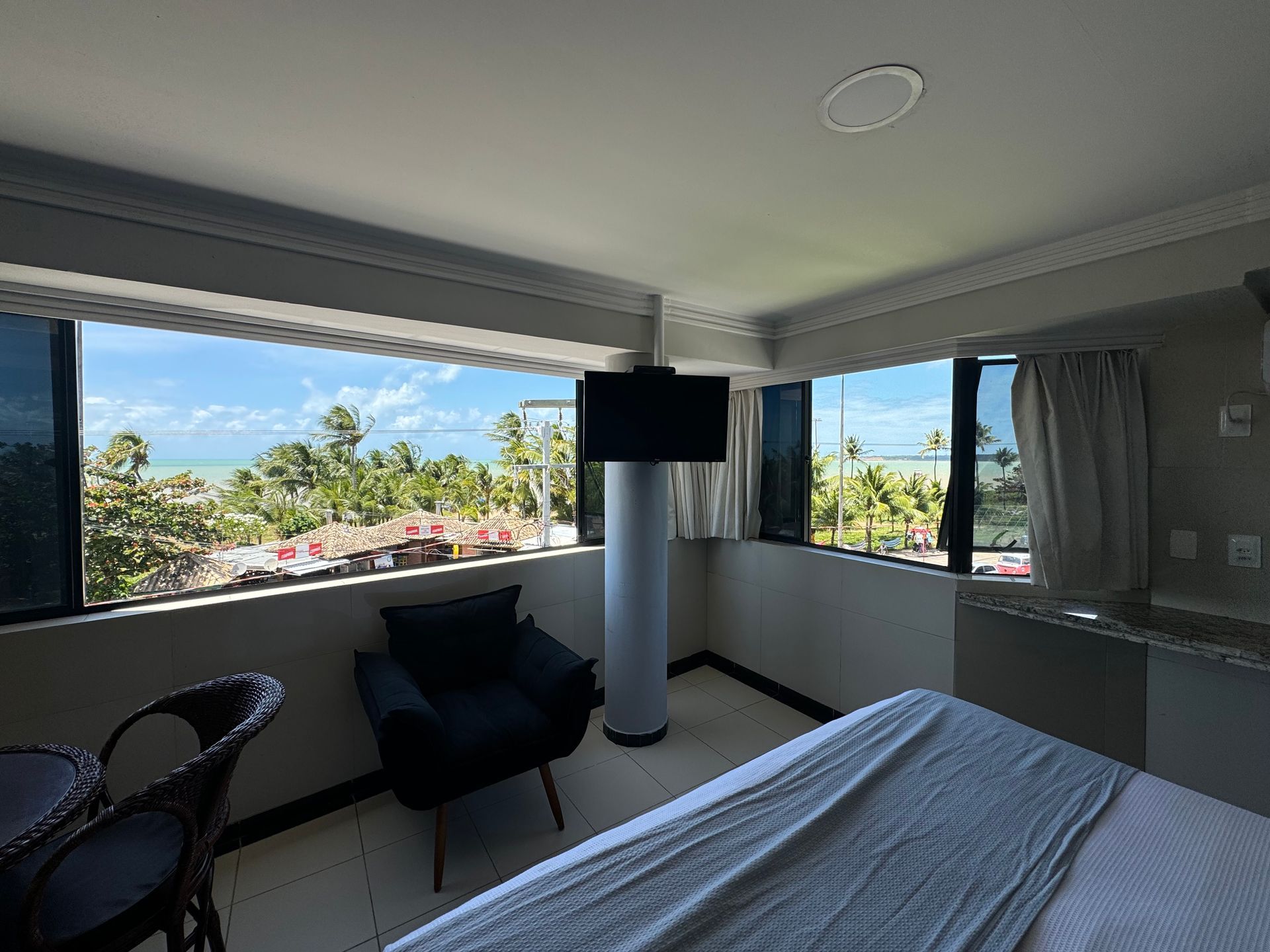 Um quarto de hotel com uma cama, uma poltrona escura e uma TV fixada na parede, com vista para uma praia com palmeiras através de grandes janelas.