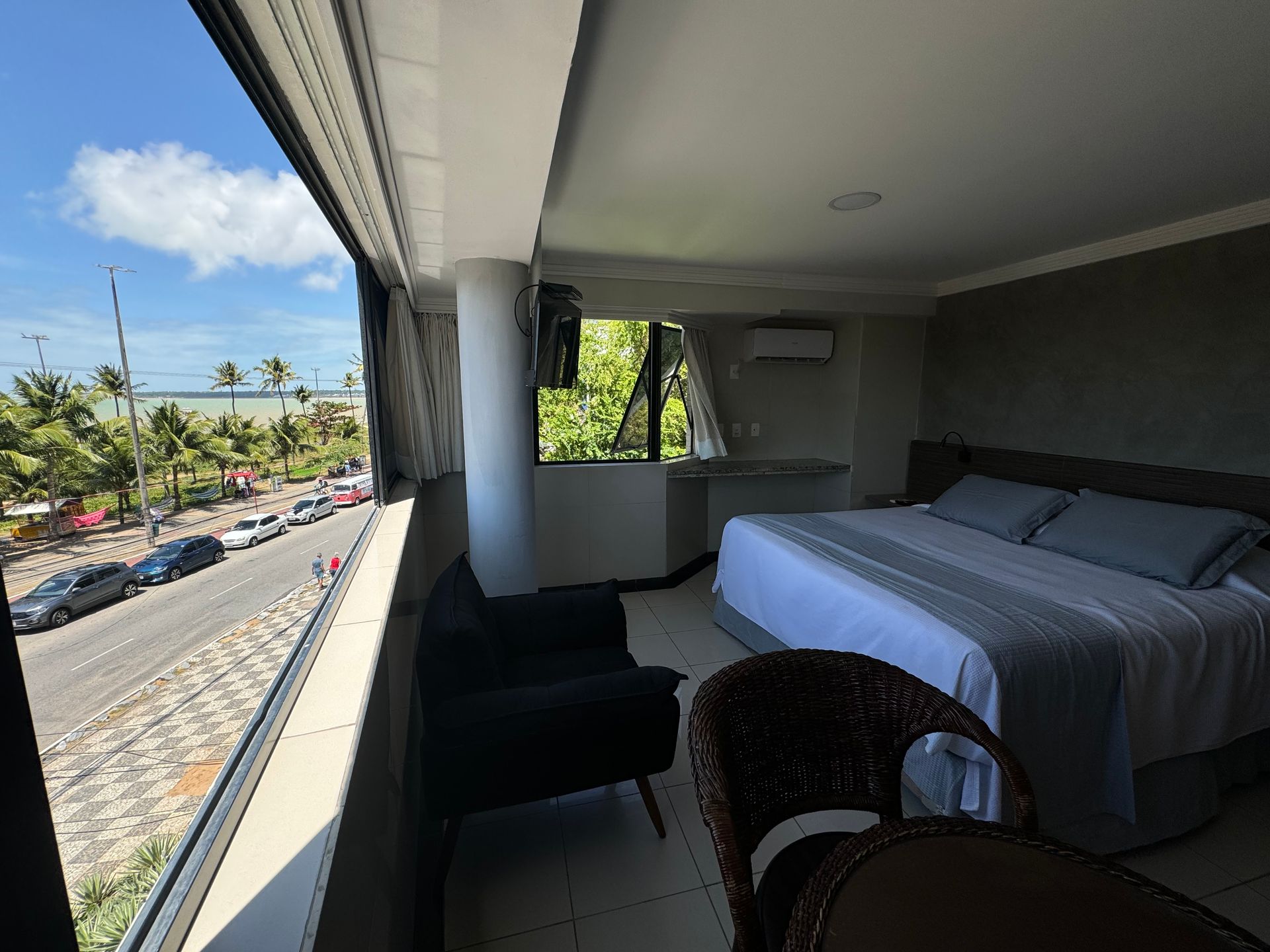Quarto de hotel com cama grande, poltrona e varanda com vista para uma avenida costeira ladeada por palmeiras e para o oceano.