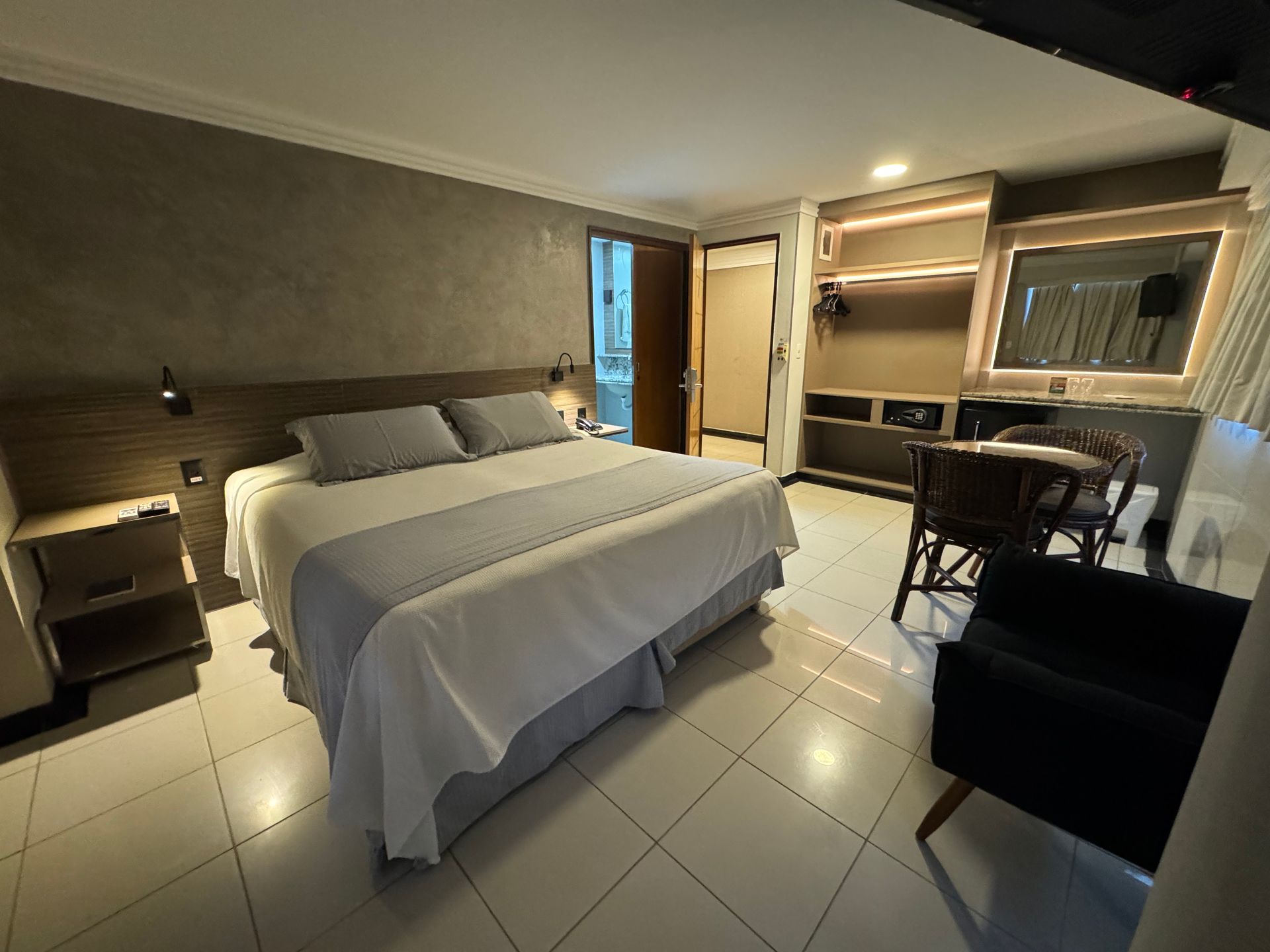 Um quarto de hotel moderno com uma cama grande com roupa de cama cinza, uma escrivaninha com cadeira e um closet aberto com piso de azulejo branco.