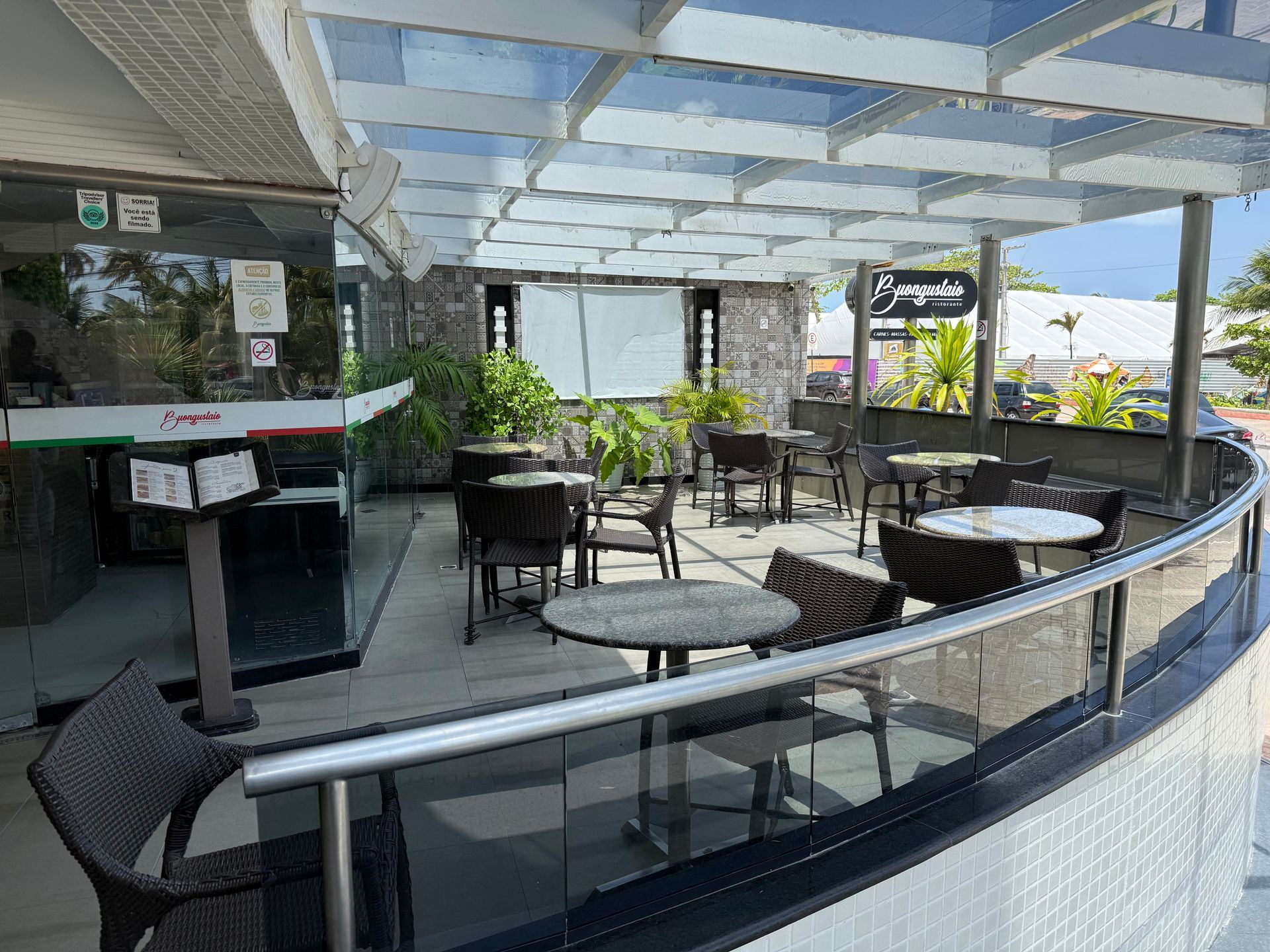 Área externa de um restaurante com várias mesas e cadeiras pequenas dispostas sob um moderno teto de pérgola branco.
