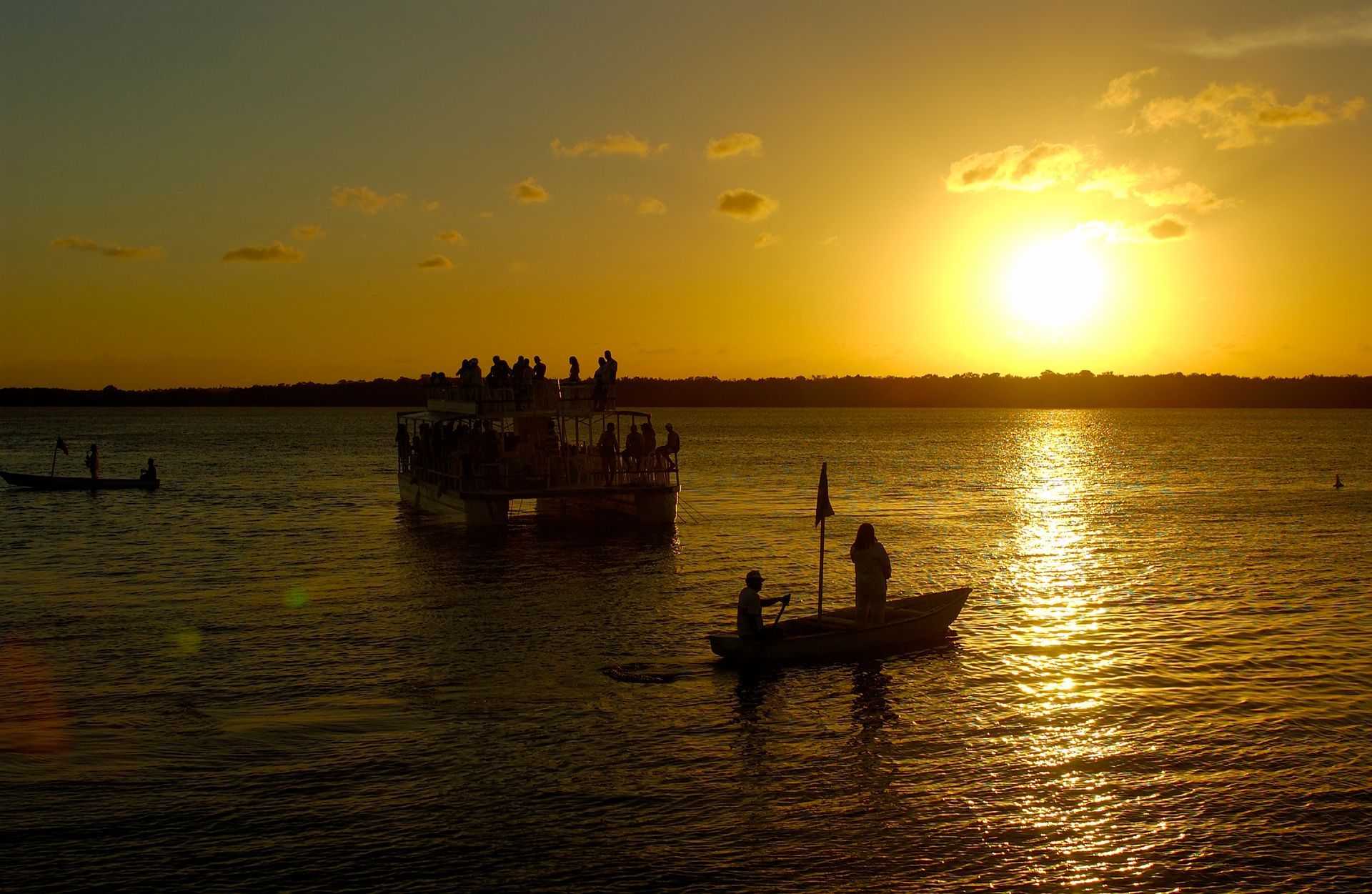 Um pôr do sol dourado sobre águas calmas, com as silhuetas de um grande barco de passageiros e dois barcos de pesca menores.