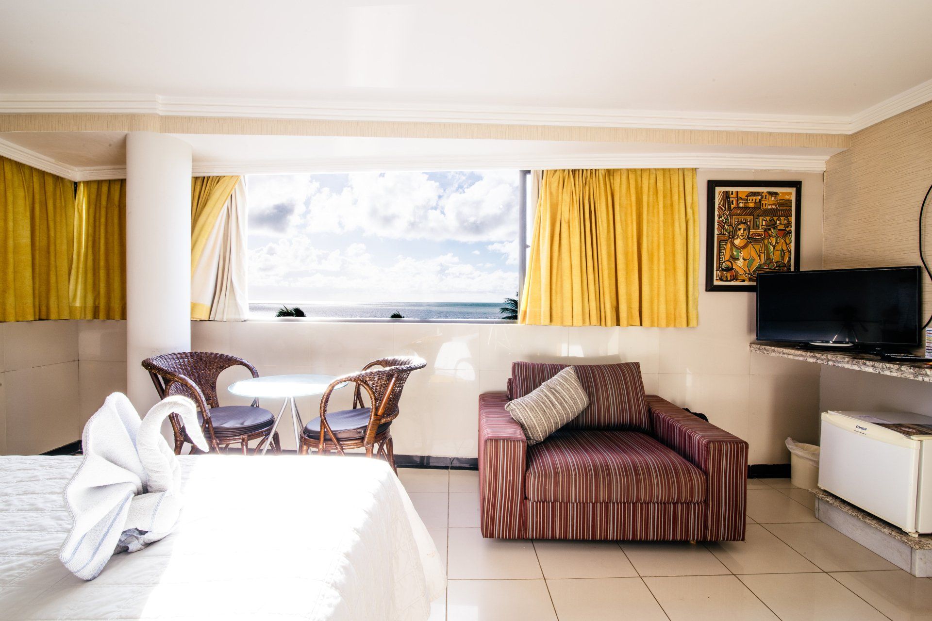 Um quarto de hotel luminoso dispõe de uma cama com uma toalha em forma de cisne, uma pequena mesa com duas cadeiras de vime, um sofá e vista para o mar.