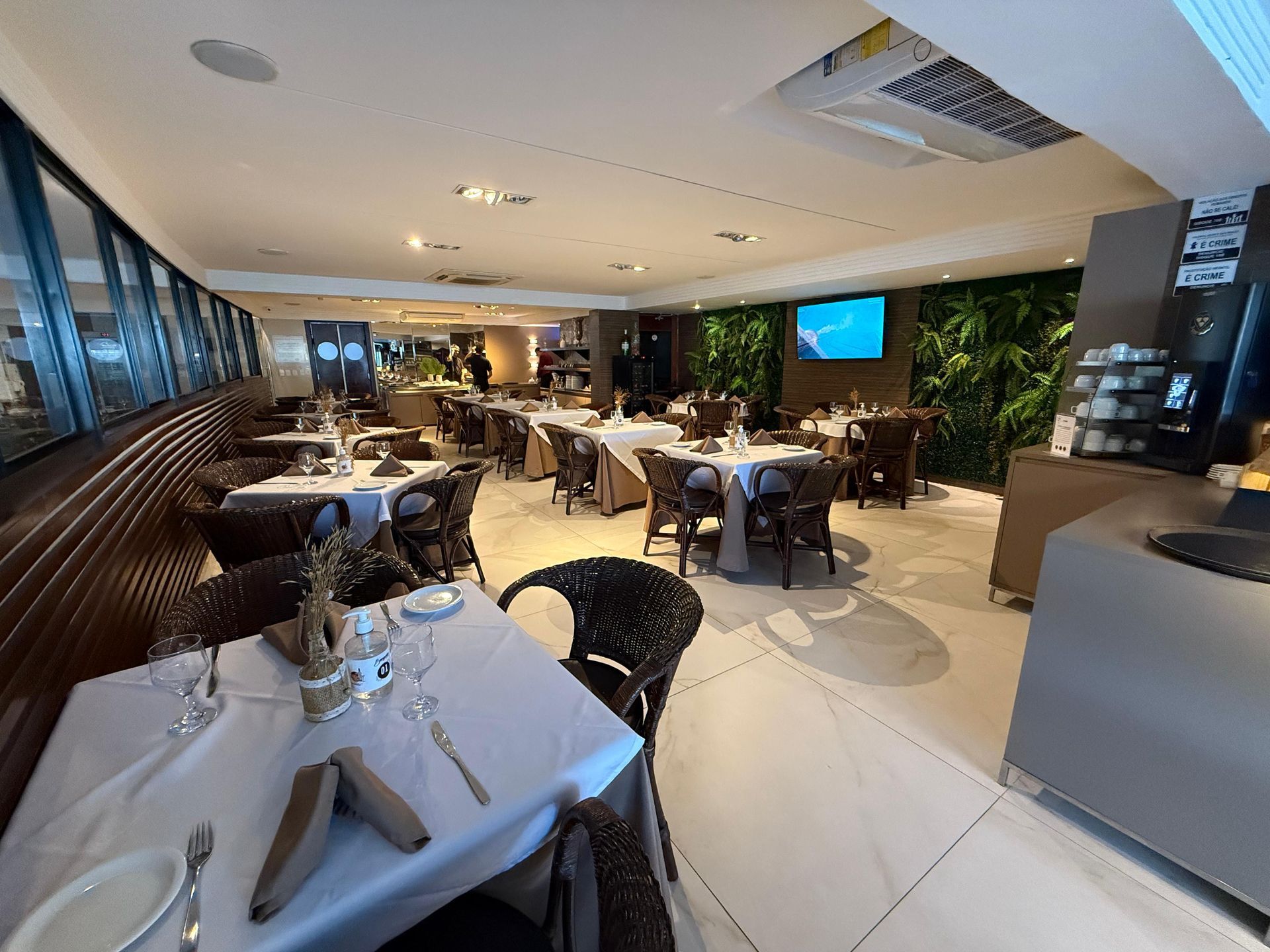 Uma sala de jantar de restaurante moderna com mesas cobertas com toalhas brancas, cadeiras de vime e uma parede com detalhes em verde, sob iluminação suave.
