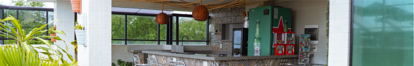 Bar externo com balcão de pedra, luminárias pendentes e vegetação, visível através de colunas arquitetônicas brancas.