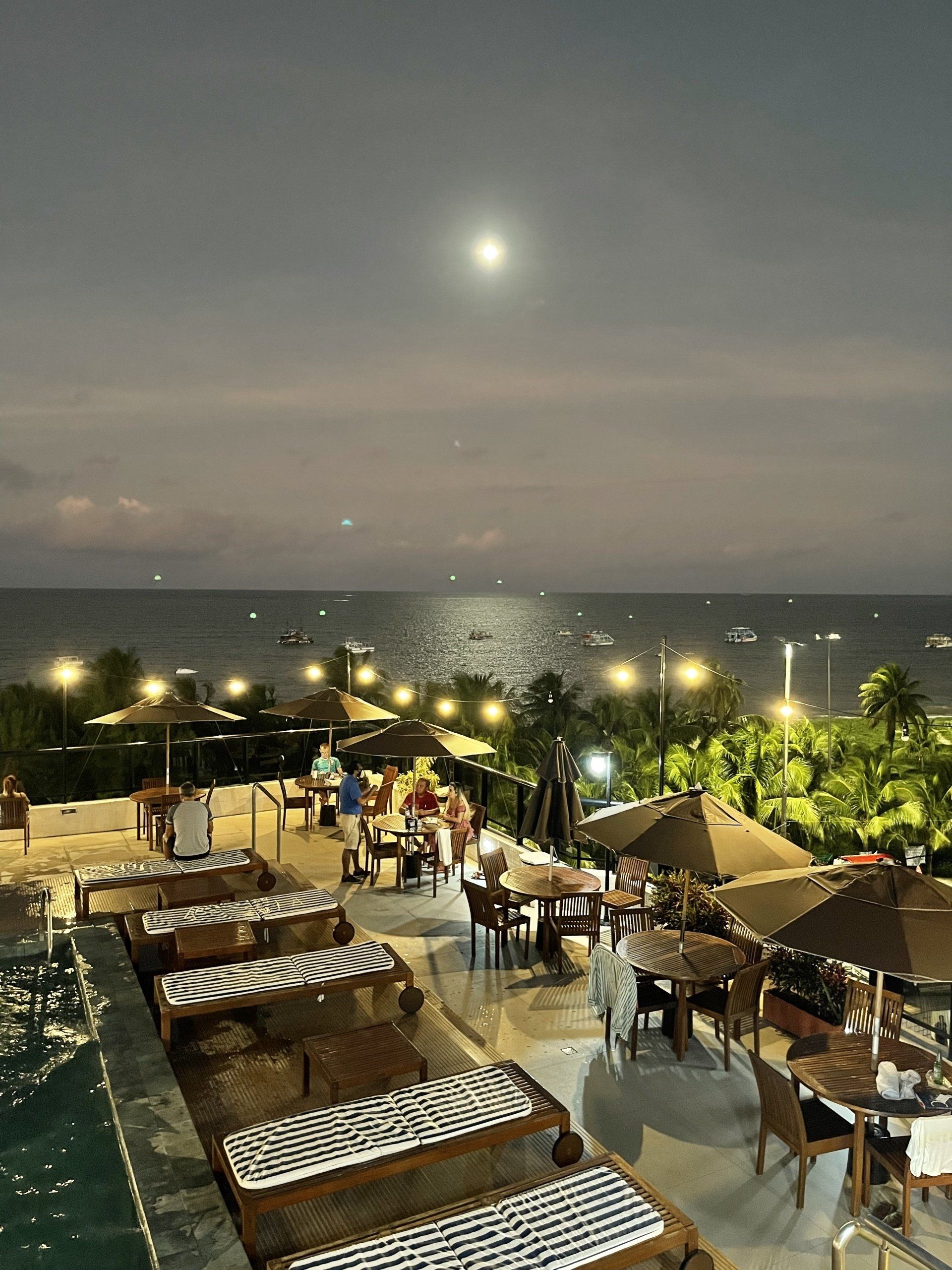 Vista noturna do terraço de um restaurante à beira-mar, com espreguiçadeiras e mesas sob cordões de luzes aconchegantes.
