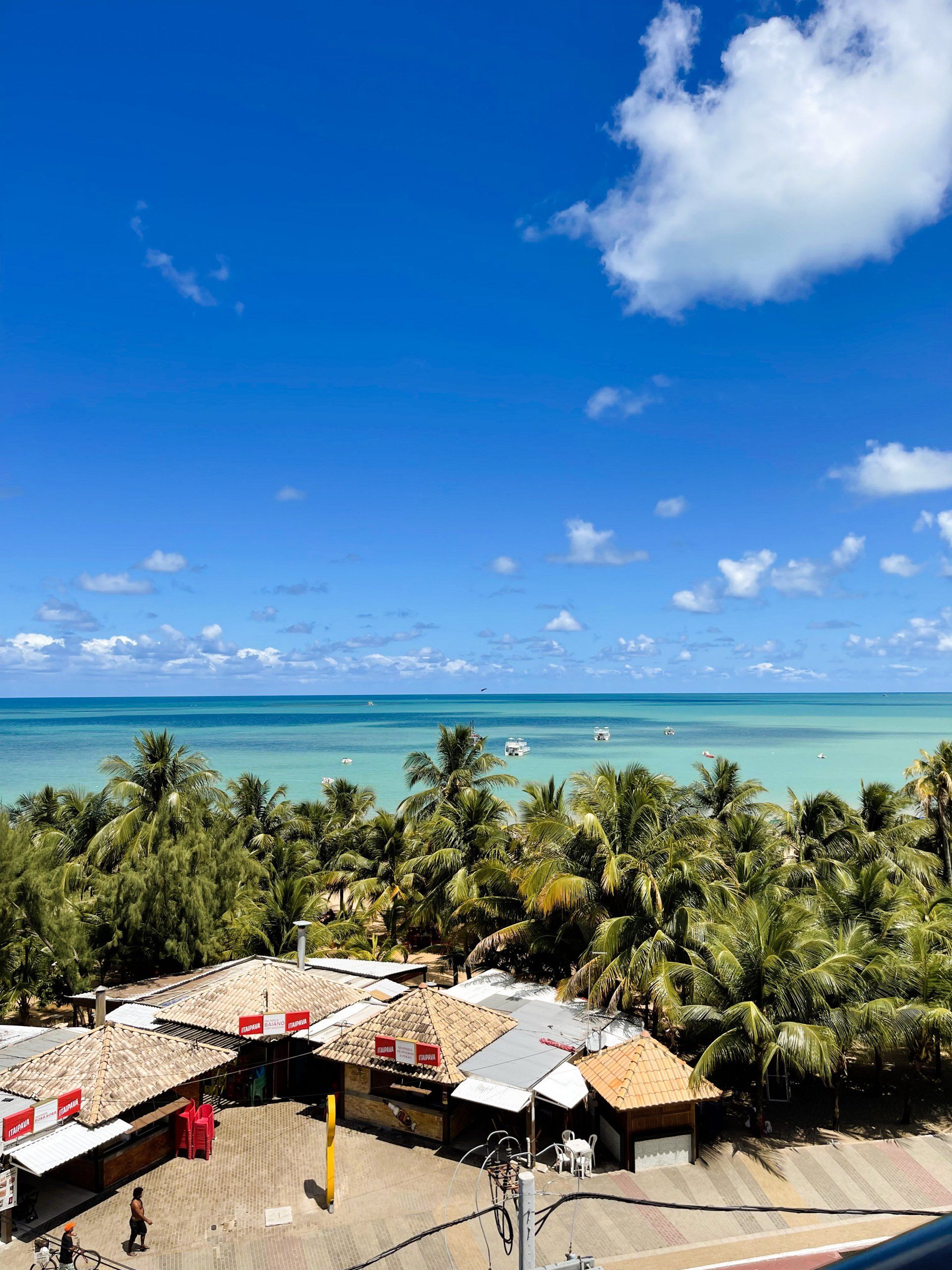 Vista de cabanas de praia tropicais aninhadas entre palmeiras, com vista para um mar calmo e turquesa sob um céu azul brilhante e ensolarado.