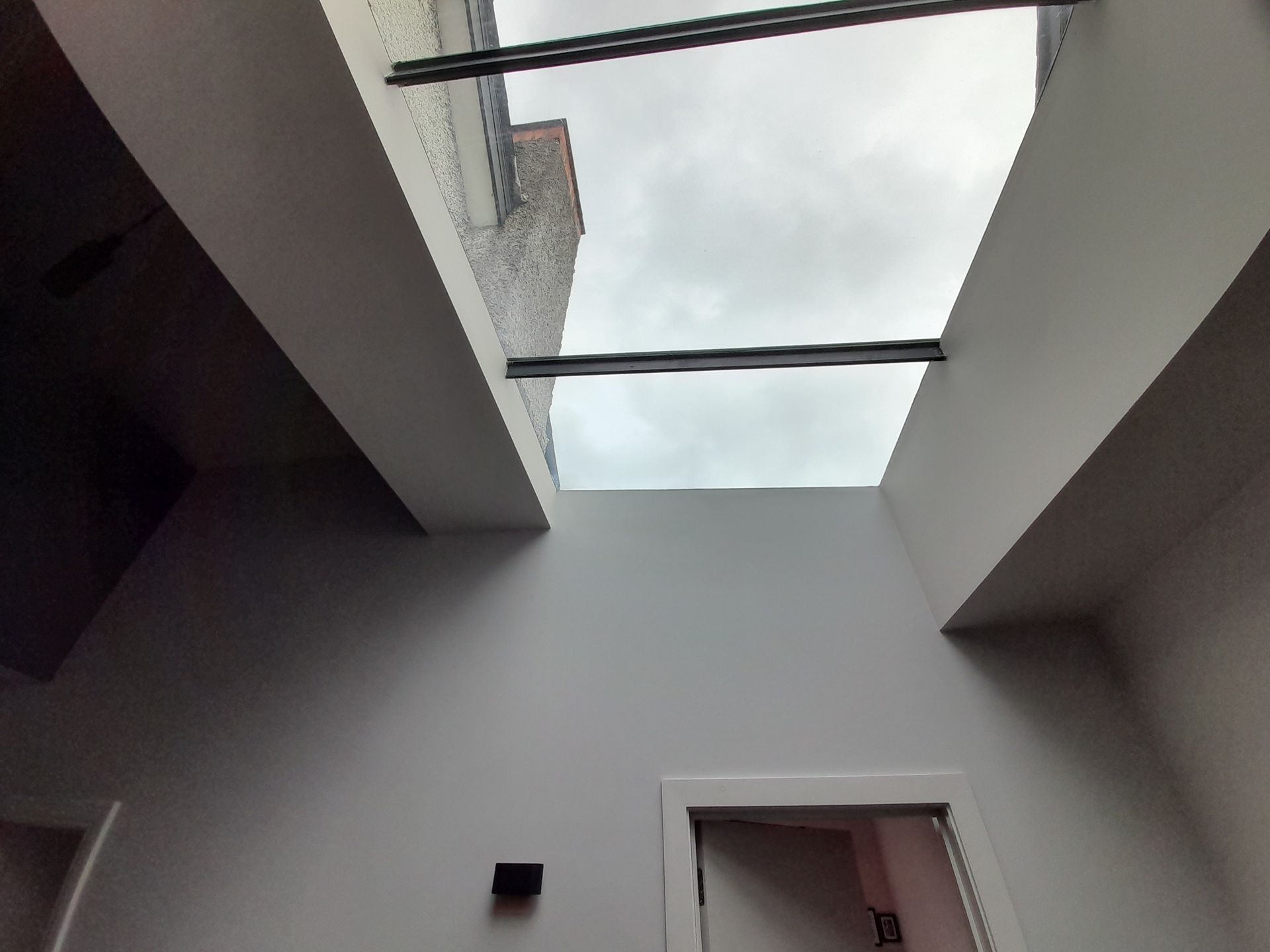 Looking up through an open skylight in a white interior ceiling with dark support beams and a wall opening below