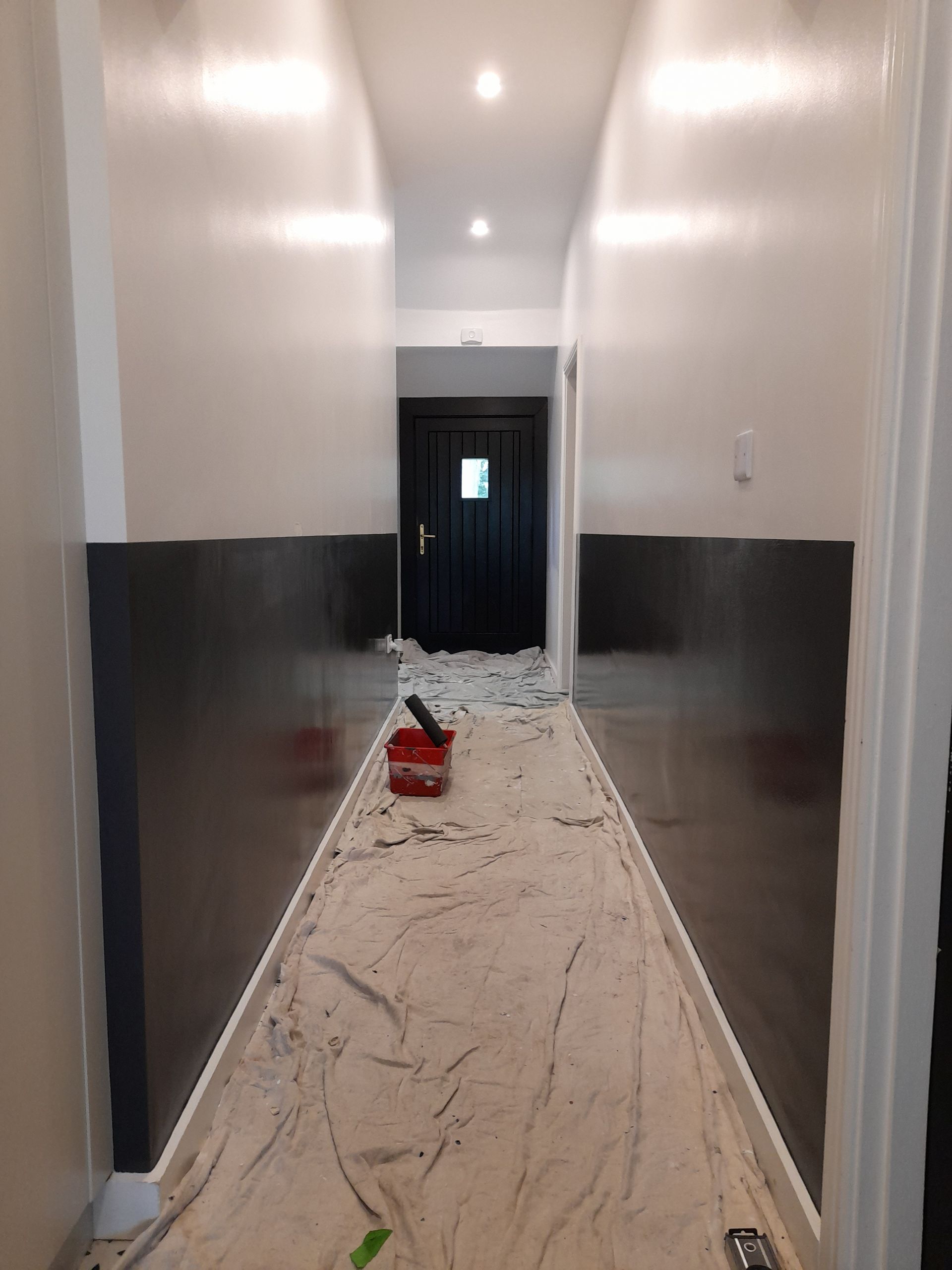 Narrow hallway with fresh plaster on the floor, dark lower walls, and a red tool bucket near the center.