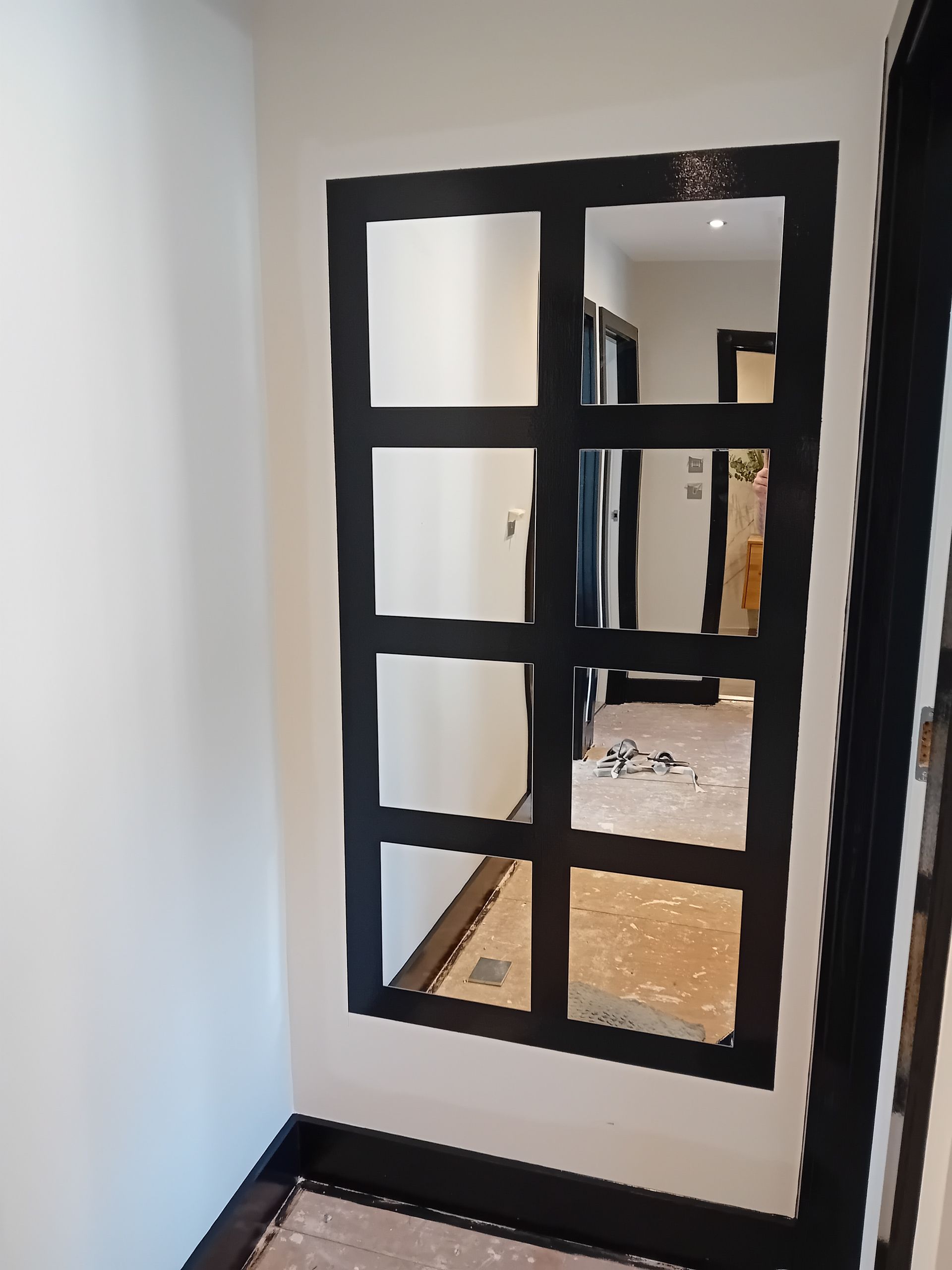 Narrow hallway with a black-framed mirror and white walls, reflecting a room with a patterned rug.