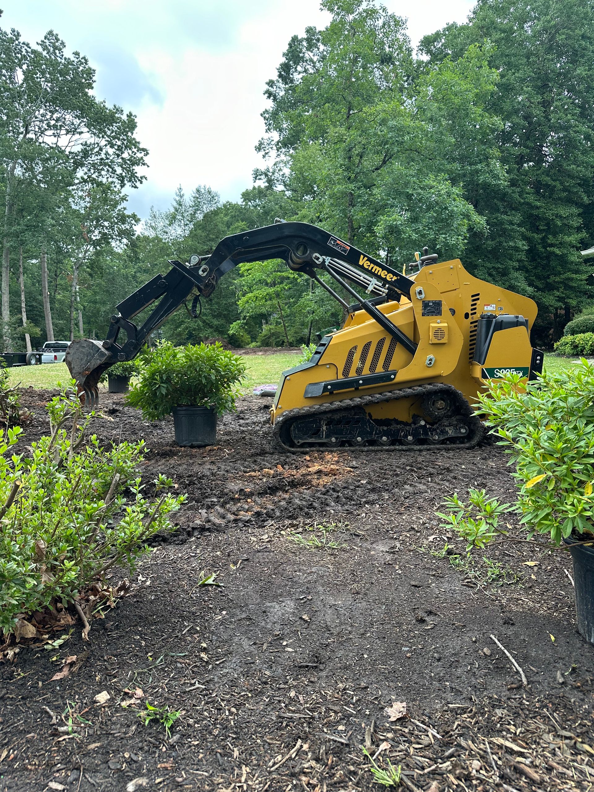 A yellow compact track loader with an extended arm and excavator attachment working in a mulched garden area.