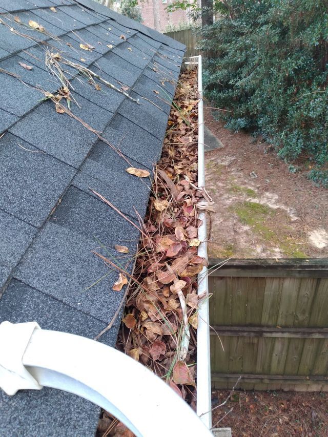 A white house gutter overflowing with dry, brown autumn leaves next to a dark gray asphalt shingle roof.