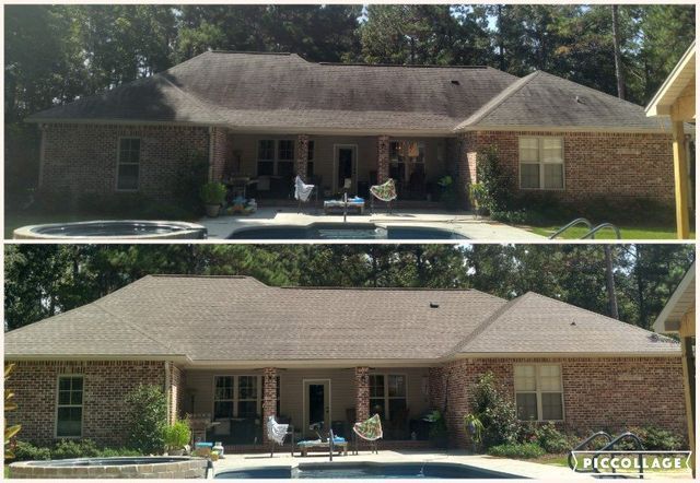 A person in work clothes uses a long pole to spray water onto the exterior of a large brick house.