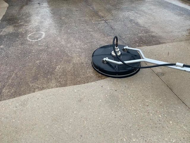 A circular pressure washer surface cleaner cleans a dirty concrete driveway, creating a clear contrast with a clean area.