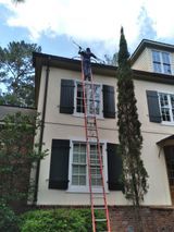 A person on an extension ladder leans against a light-colored, two-story house to clean the gutters.