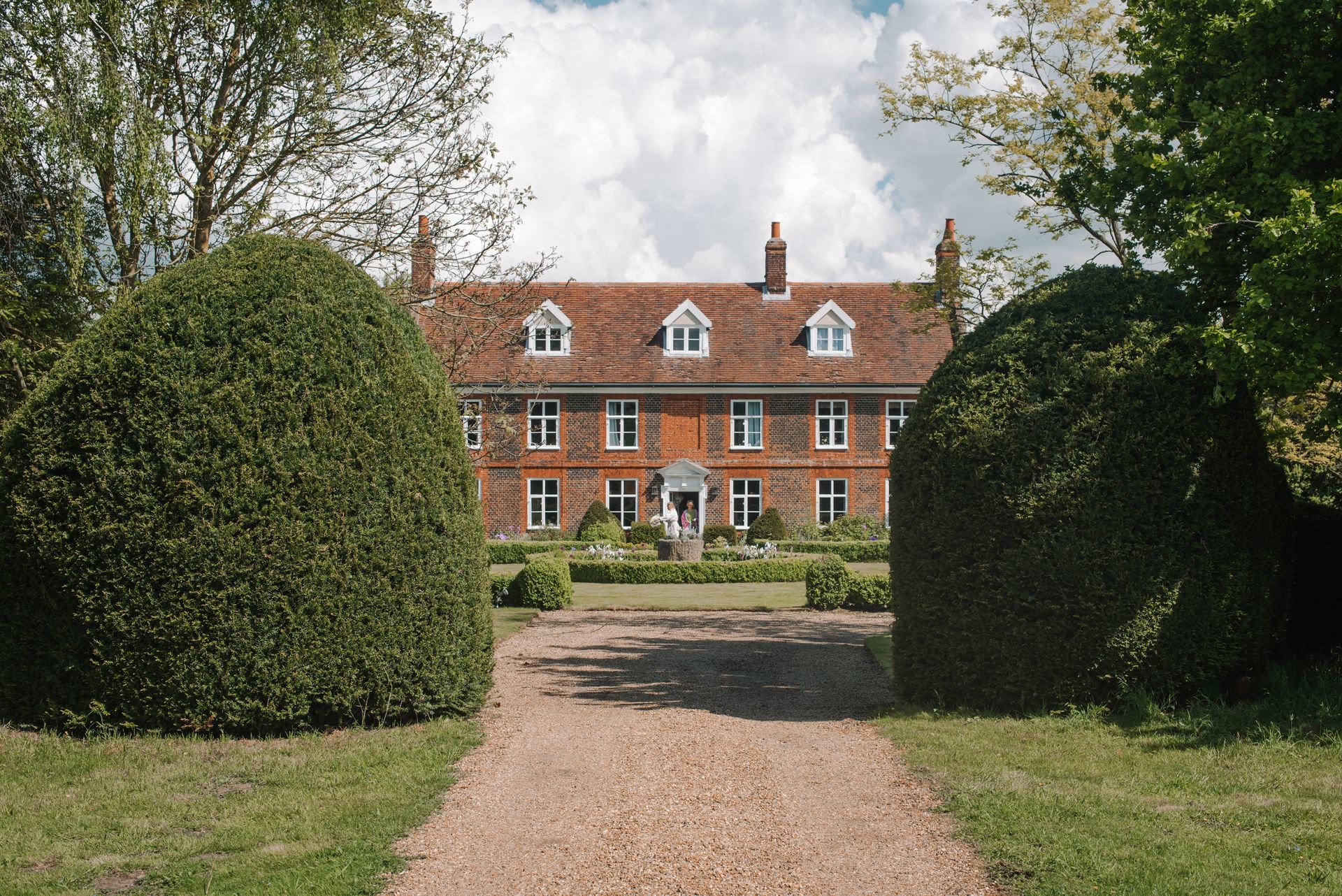 Gravel path leading to a red-brick manor house framed by large hedges and trees
