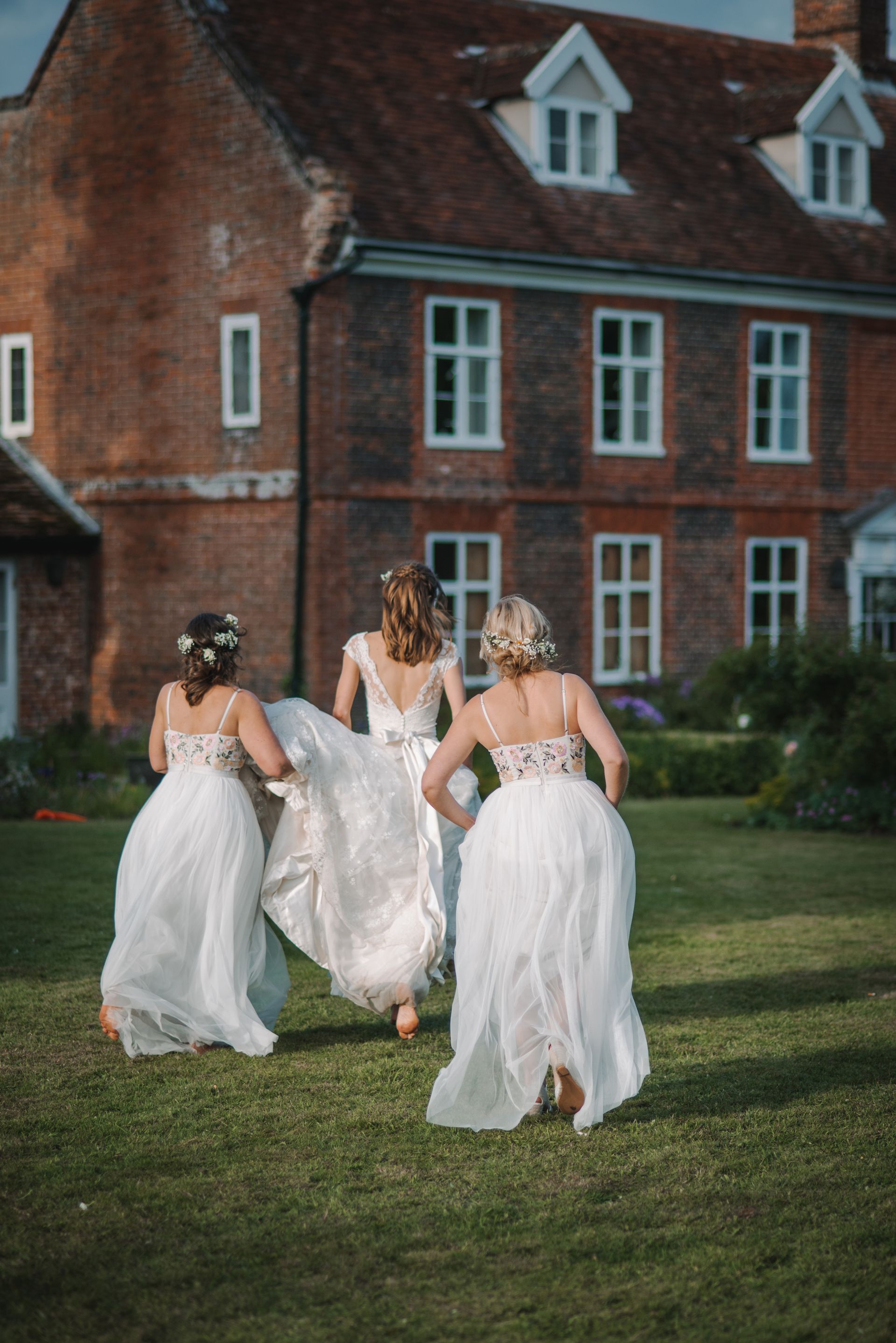 Three people in white dresses walking across a lawn toward a red brick house.
