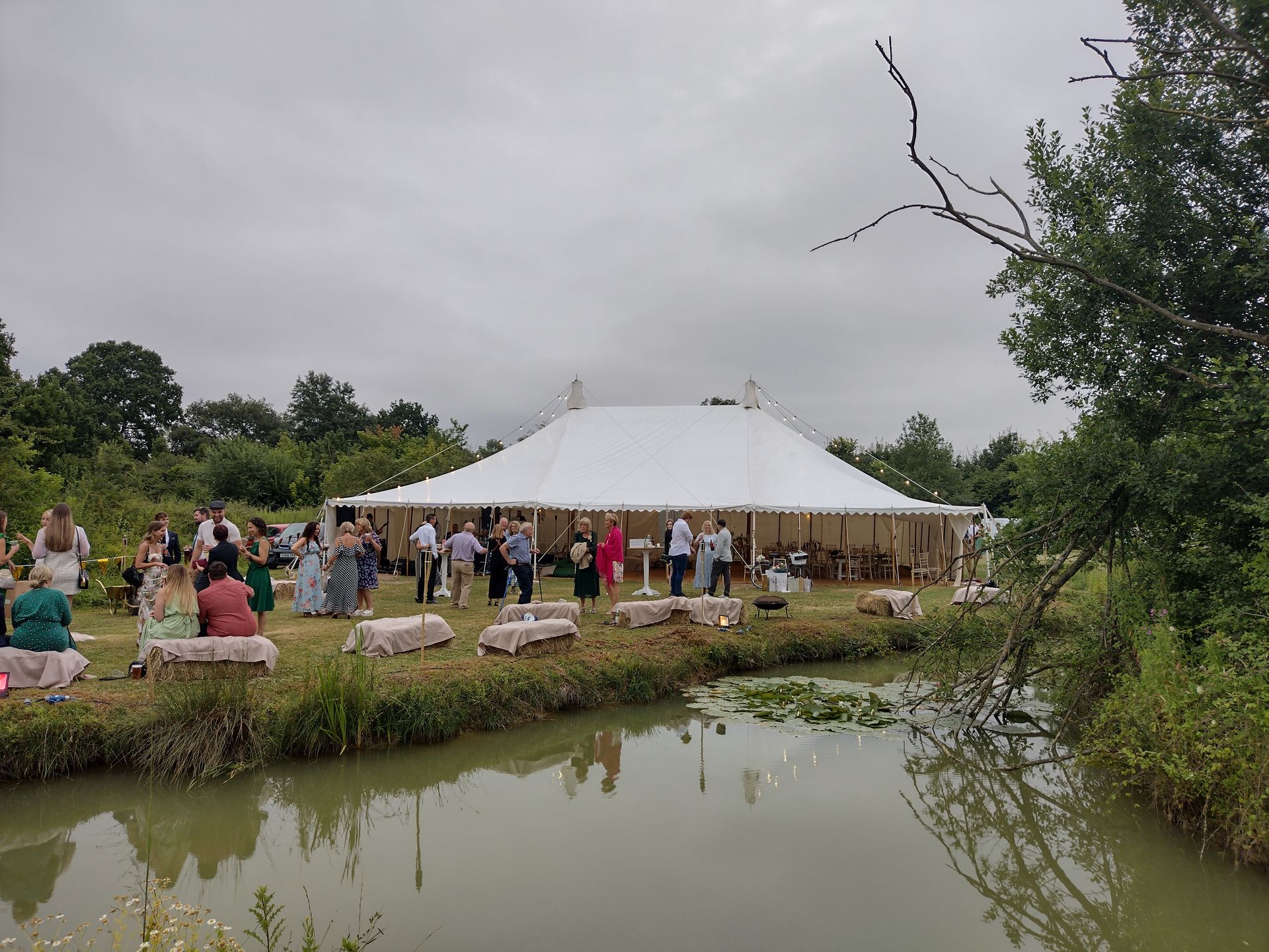 Crowd gathered beside a white event tent by a pond under a cloudy sky