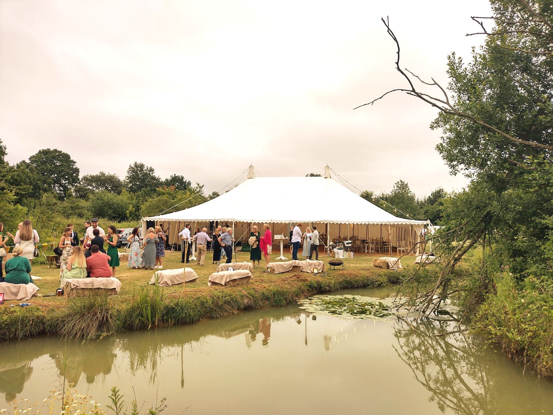 Outdoor gathering beside a pond, with a large white tent and people mingling on a grassy bank.