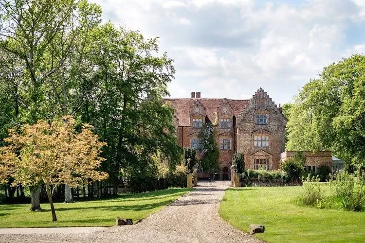 Historic brick manor house with a gravel path, green lawn, and trees in a sunny garden