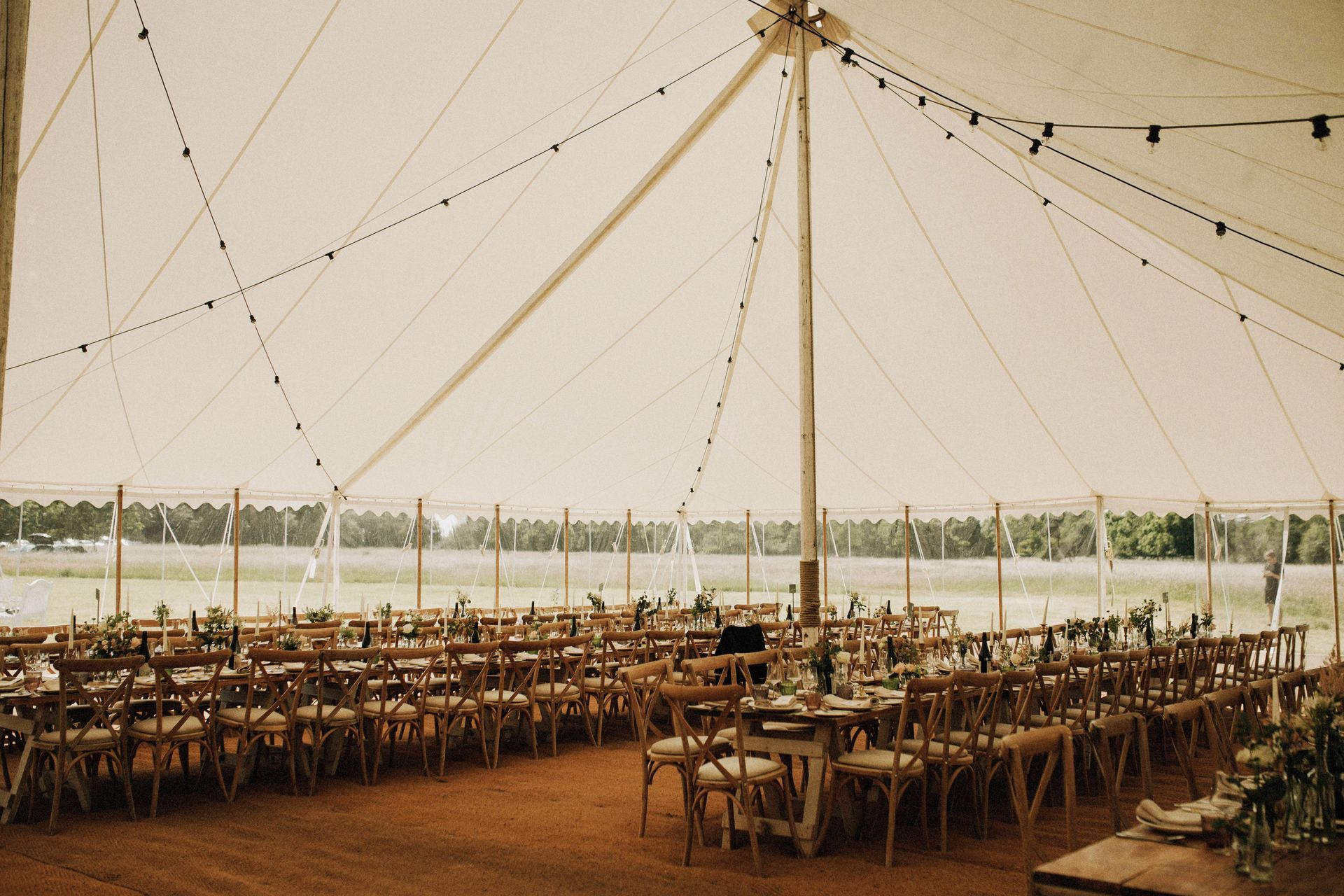 Large tented outdoor event with rows of wooden tables and chairs on a dirt floor by the water