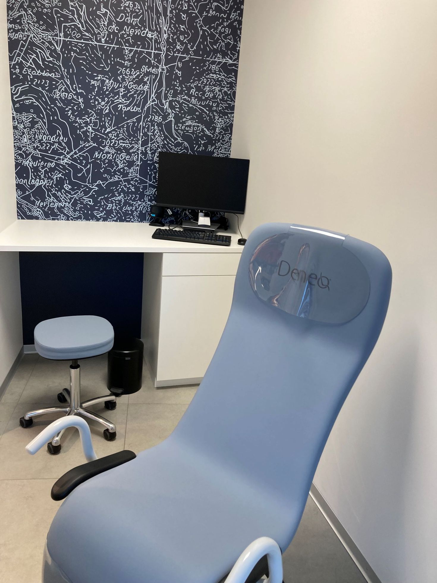 A medical exam room featuring a light blue procedure chair in the foreground and a desk with a monitor in the background.