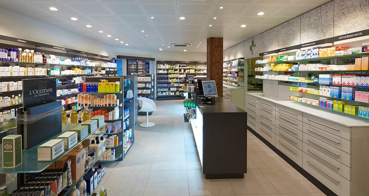 A brightly lit pharmacy interior with shelves stocked with products, a central service counter, and white tiled floors.