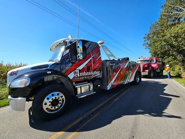 Tow truck with Ambassador logo towing a red vehicle on a sunny road.