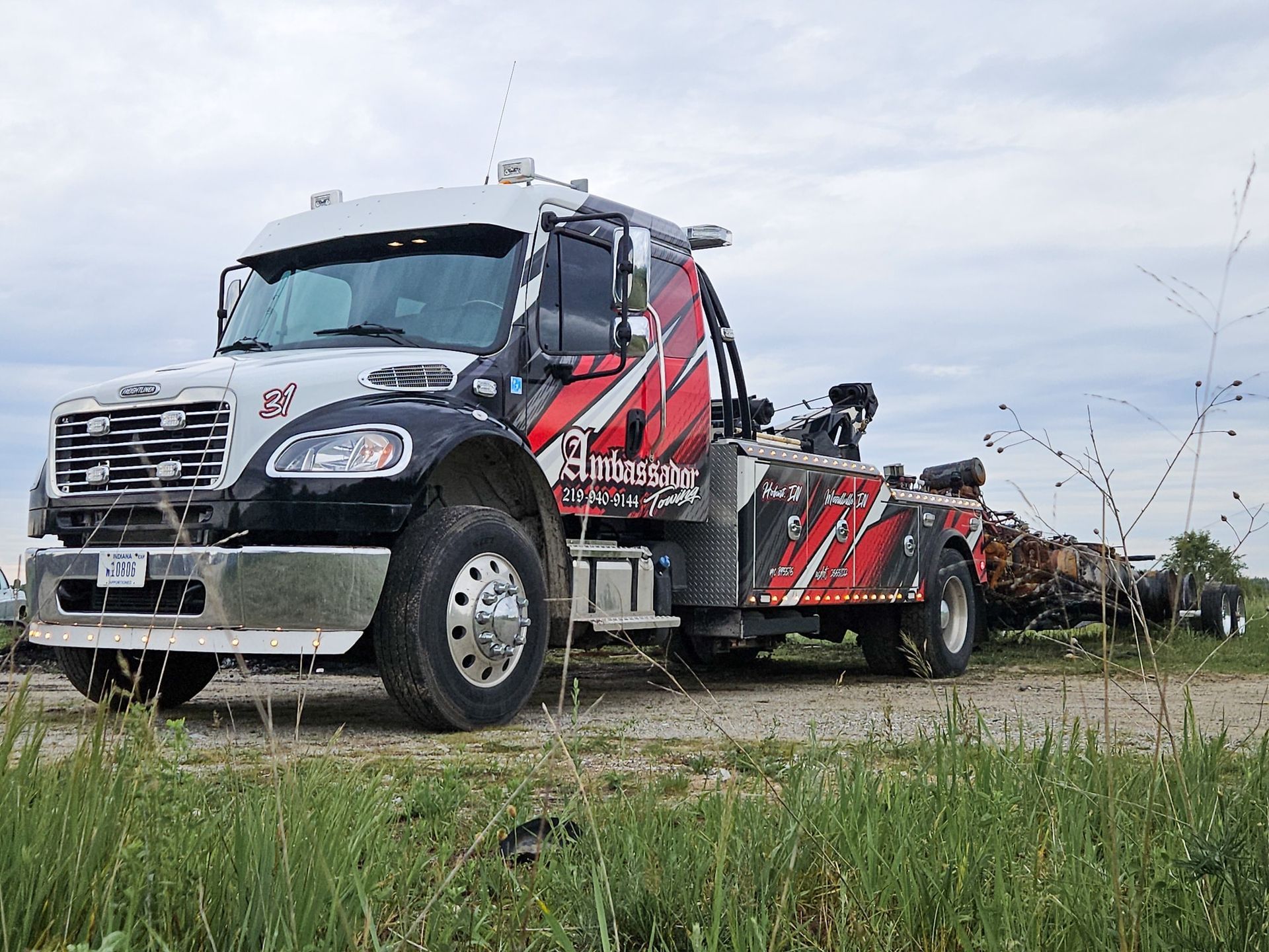 Tow truck, black and red, towing a trailer with logs on a gravel road. Cloudy sky.
