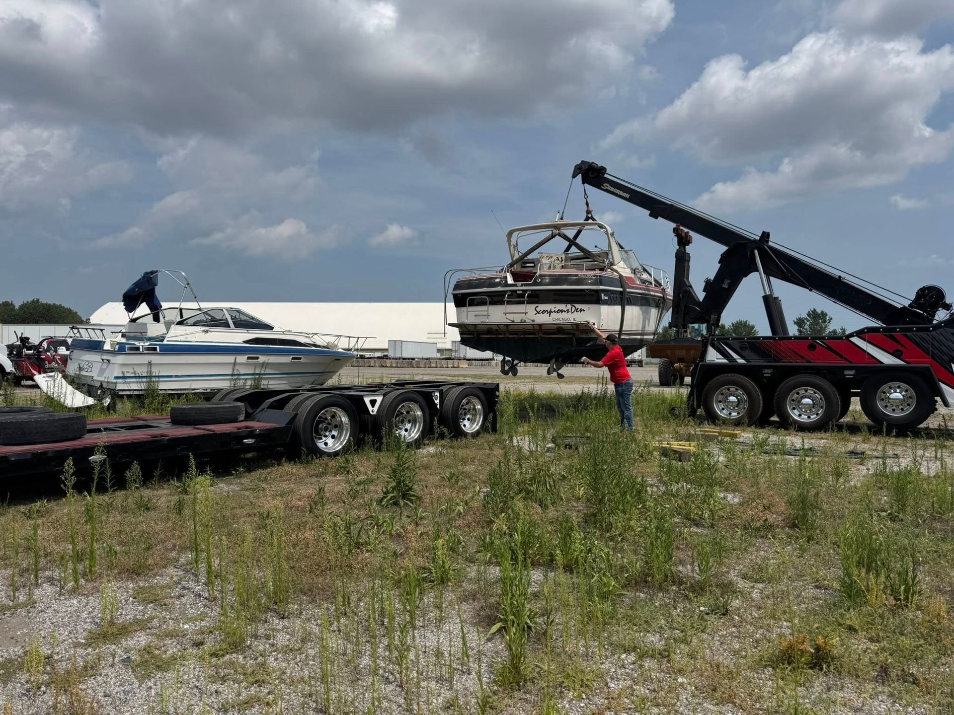 Tow truck lifting a boat, preparing to load it onto a trailer. Another boat is already on the trailer, outdoor setting.