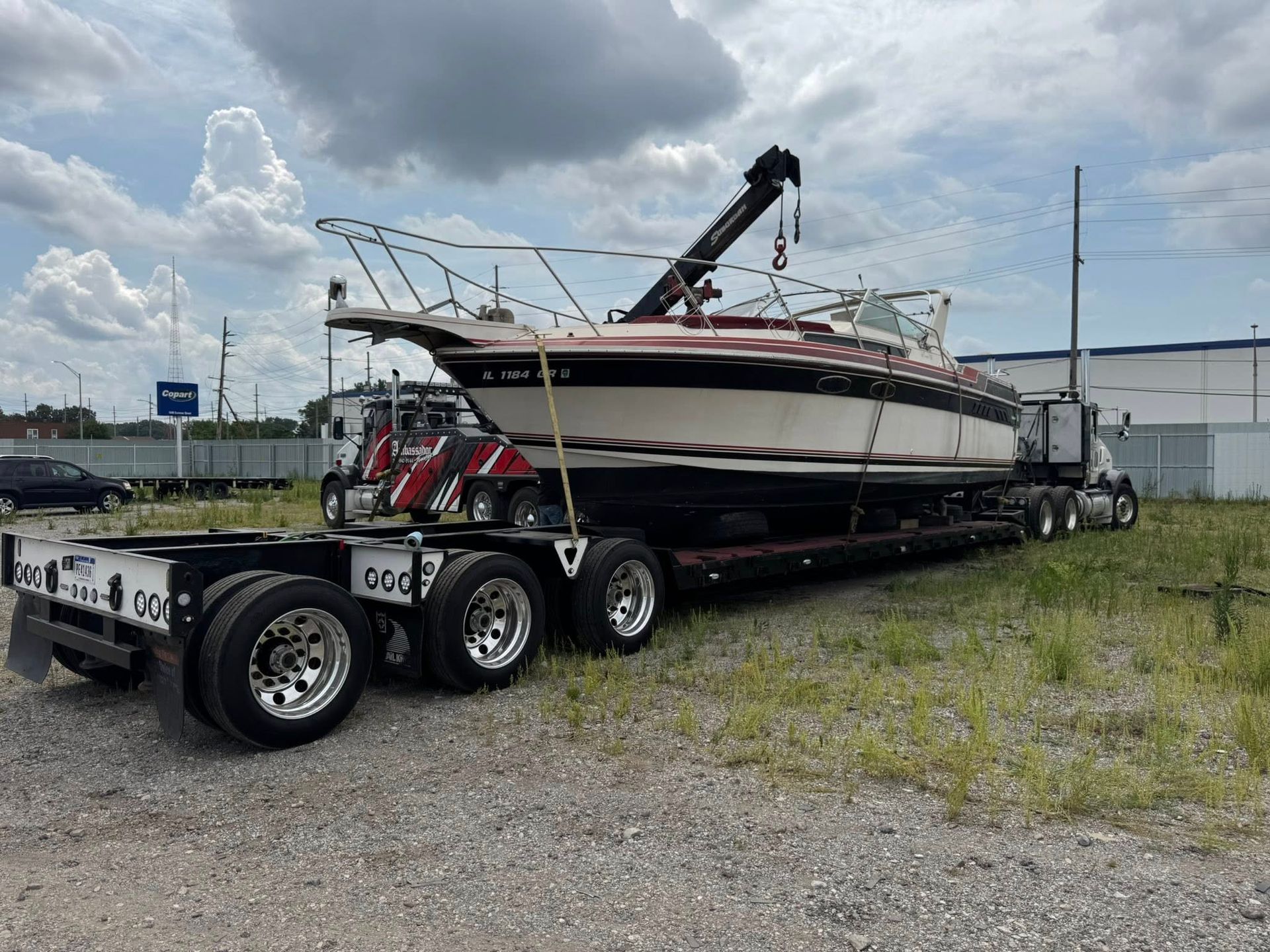 Boat on a flatbed truck, being transported on a grassy lot, under a cloudy sky.