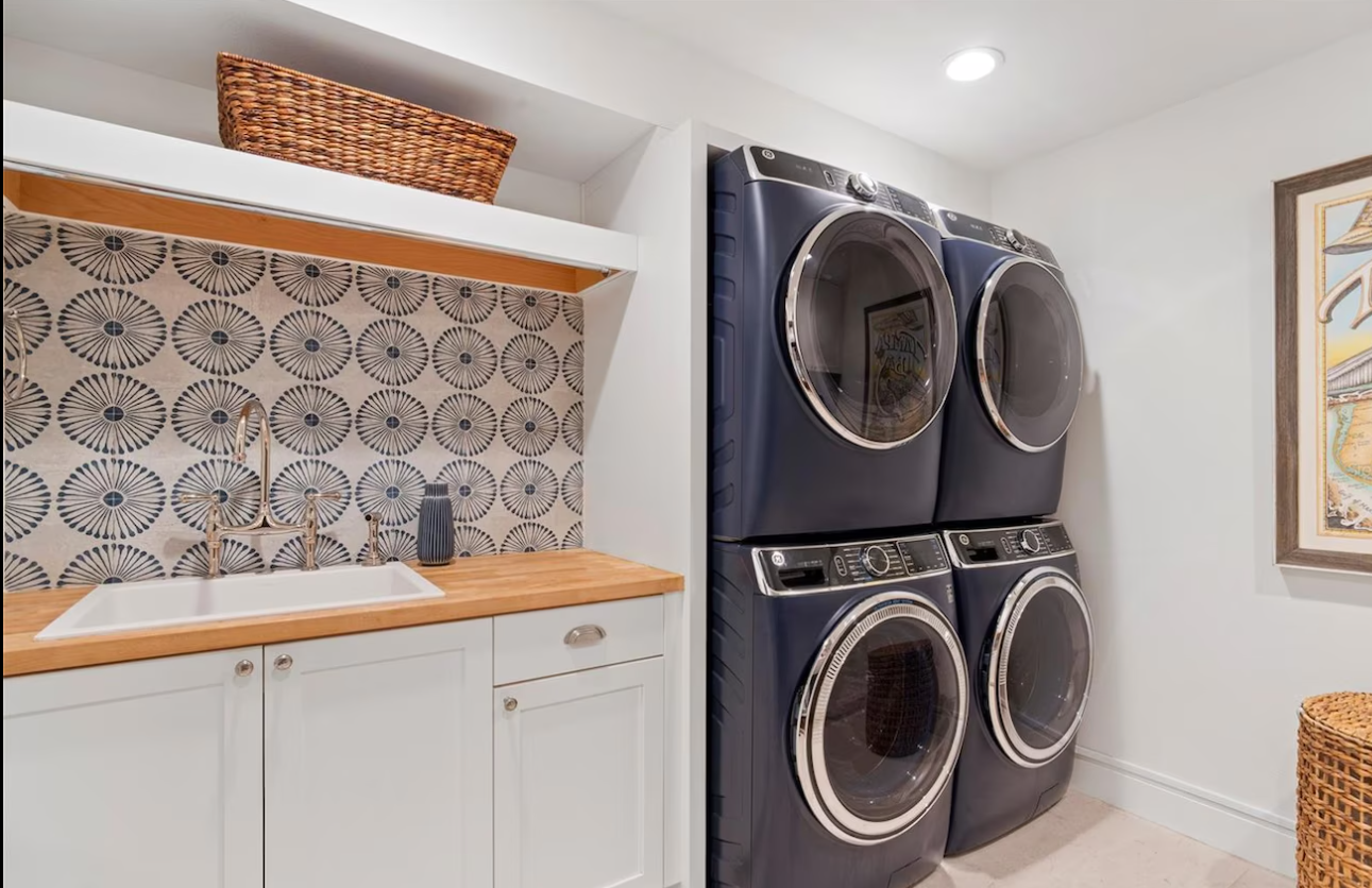 Laundry room with stacked washer-dryers, patterned backsplash, and white cabinets with wood shelves