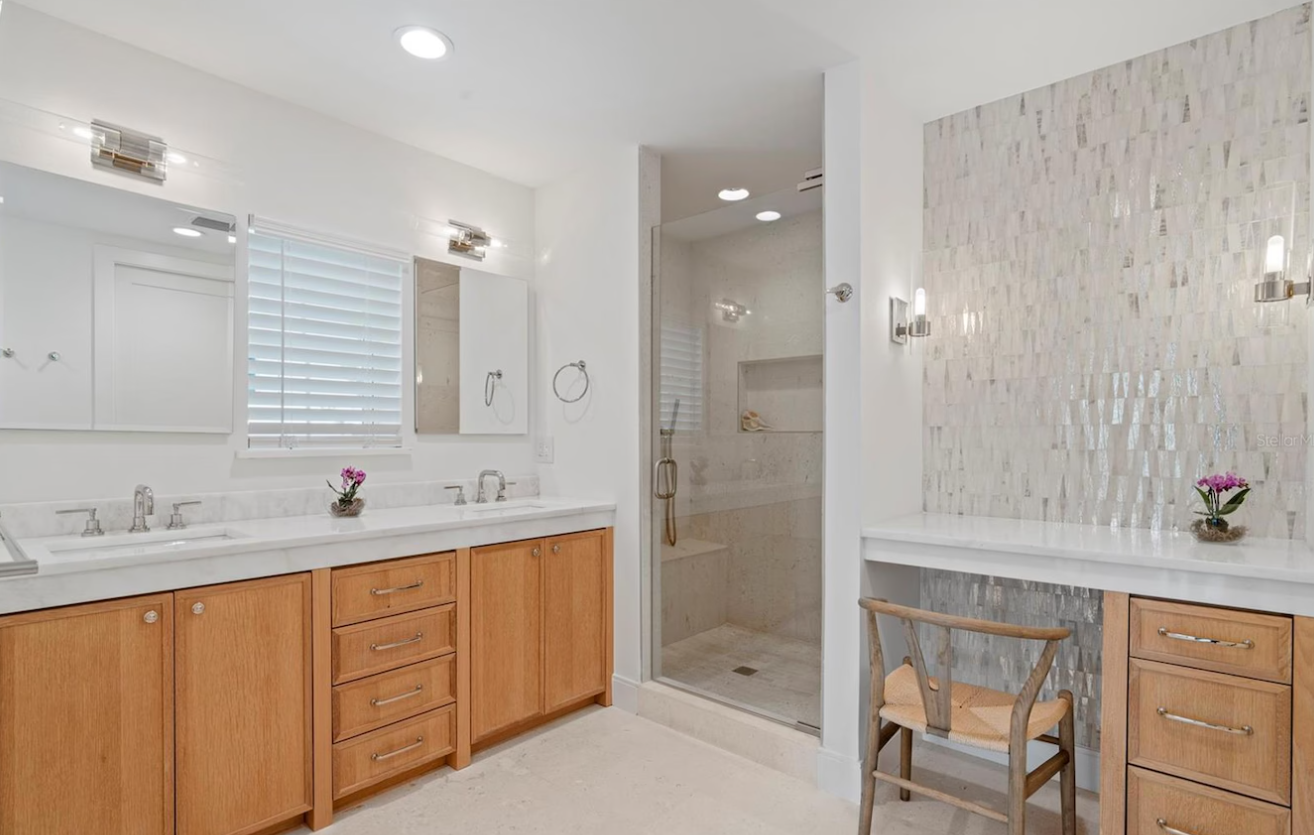 Bright modern bathroom with double vanity, large mirror, glass shower, and a small desk area