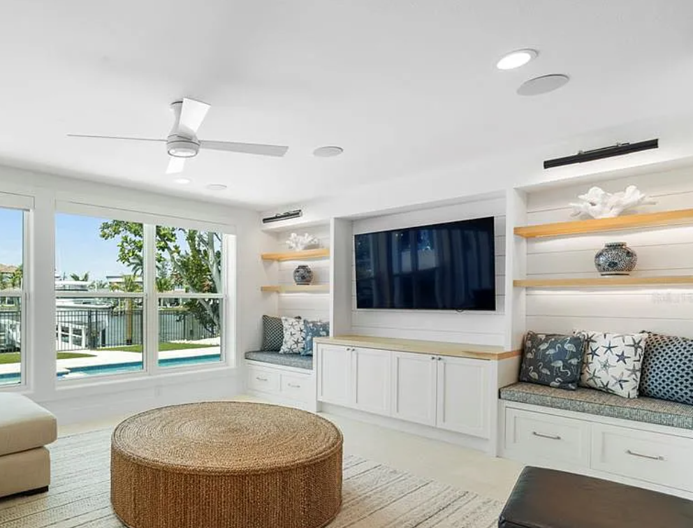 Bright white living room with wall-mounted TV, built-in bench, round ottoman, and sliding doors to a pool view