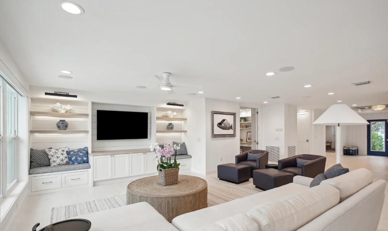 Bright modern living room with white sofas, wall-mounted TV, wicker table, and dark armchairs.