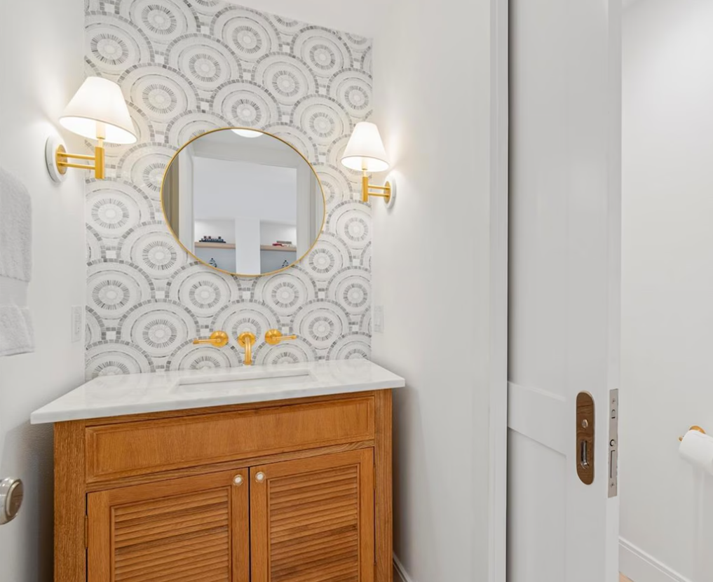 Bathroom vanity with wooden cabinet, round mirror, brass sconces, and patterned wallpaper