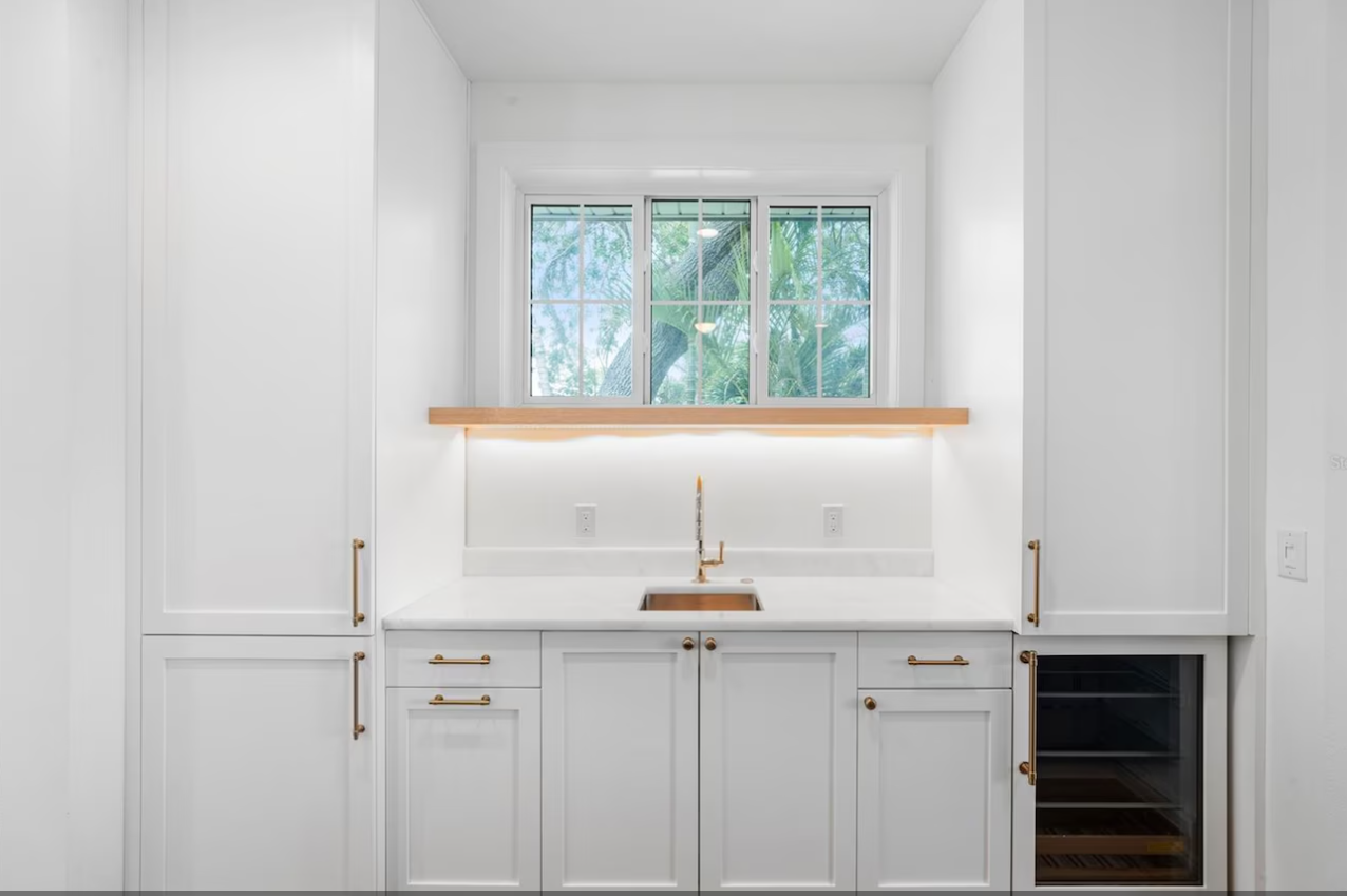 Minimal white kitchen sink area with wood shelf, brass faucet, and window above