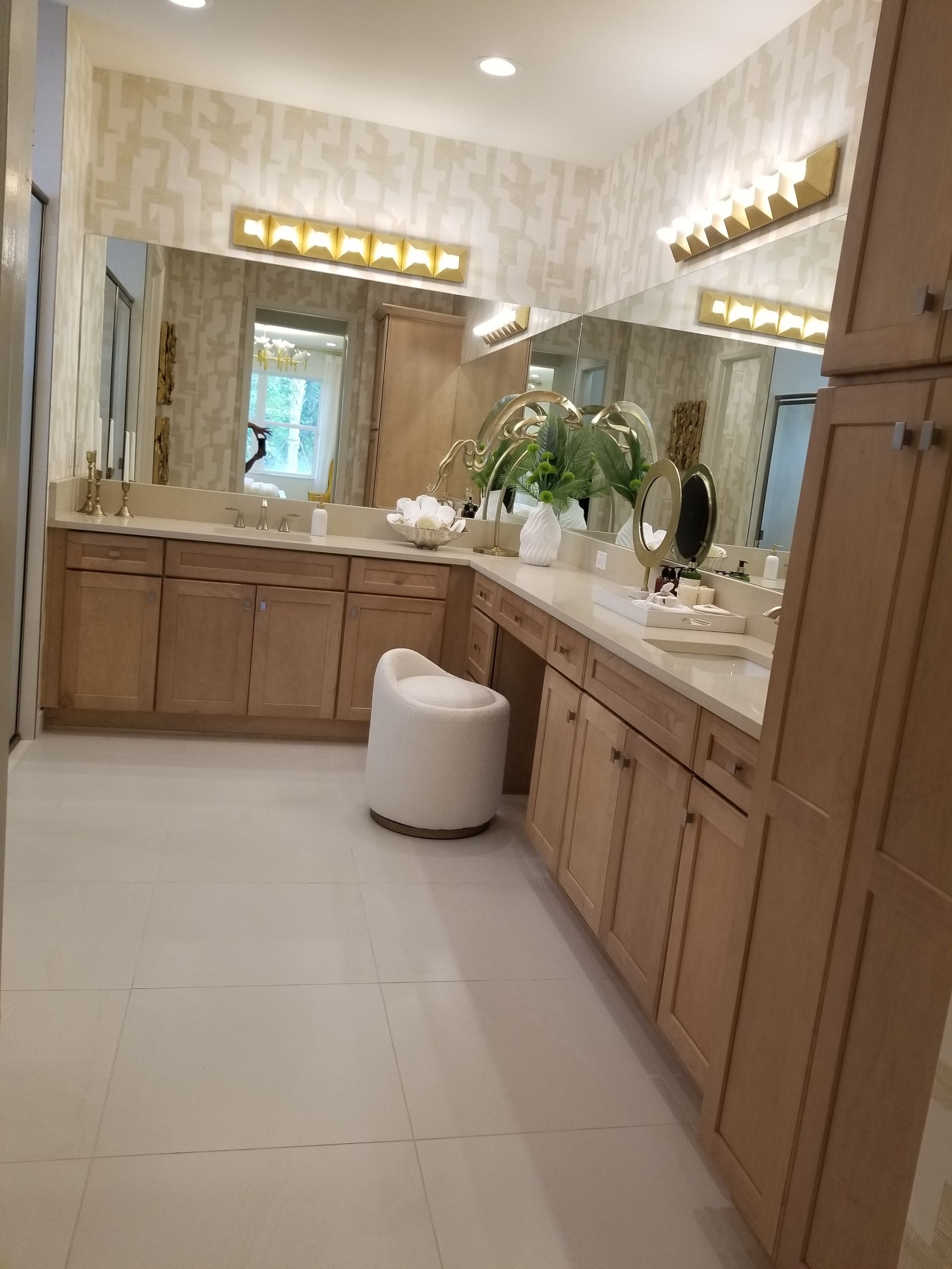 Spacious beige bathroom with wood vanities, large mirrors, wall sconces, and a white ottoman center.