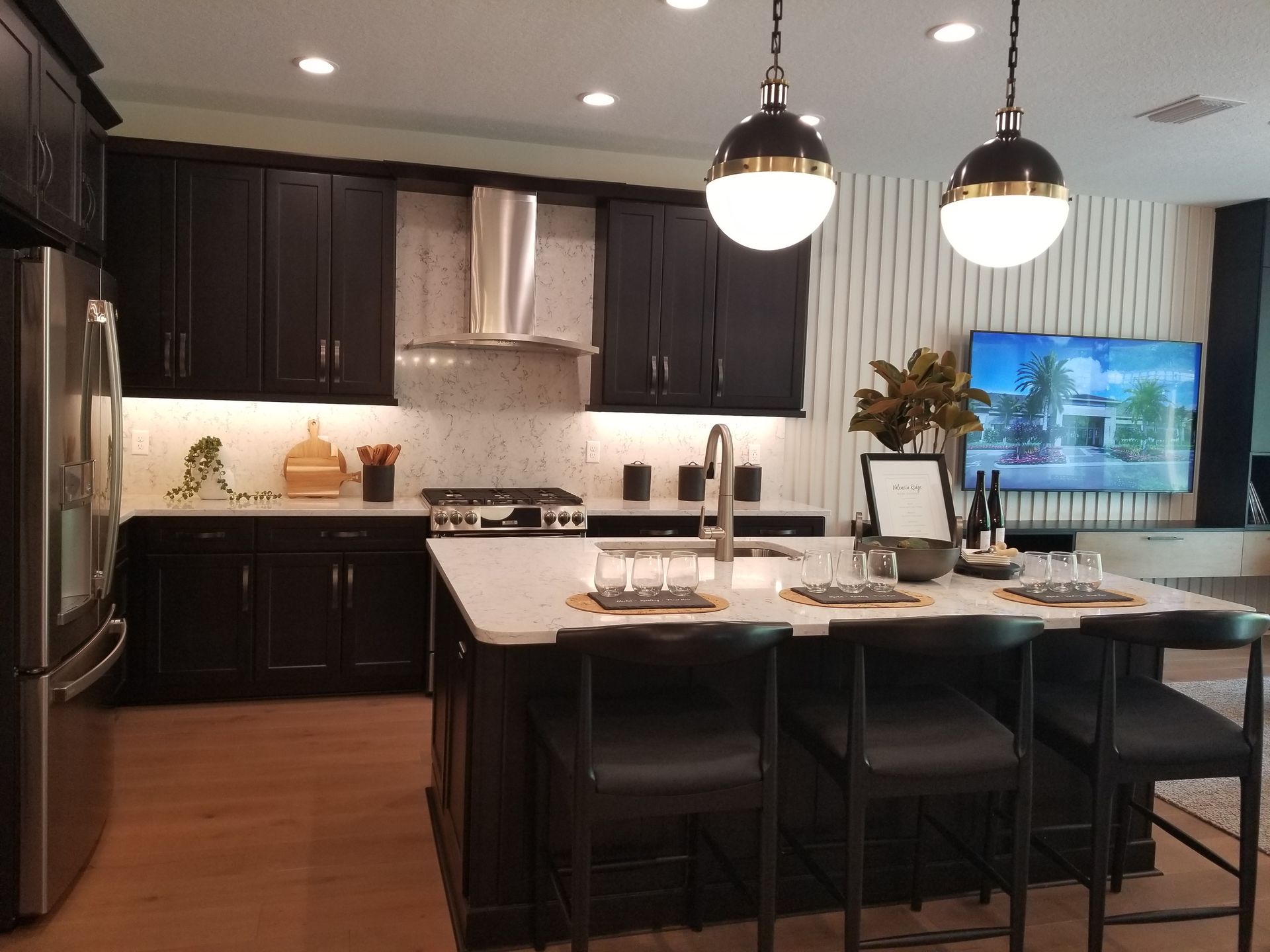 Modern kitchen with dark cabinets, island seating, pendant lights, and a TV in the background
