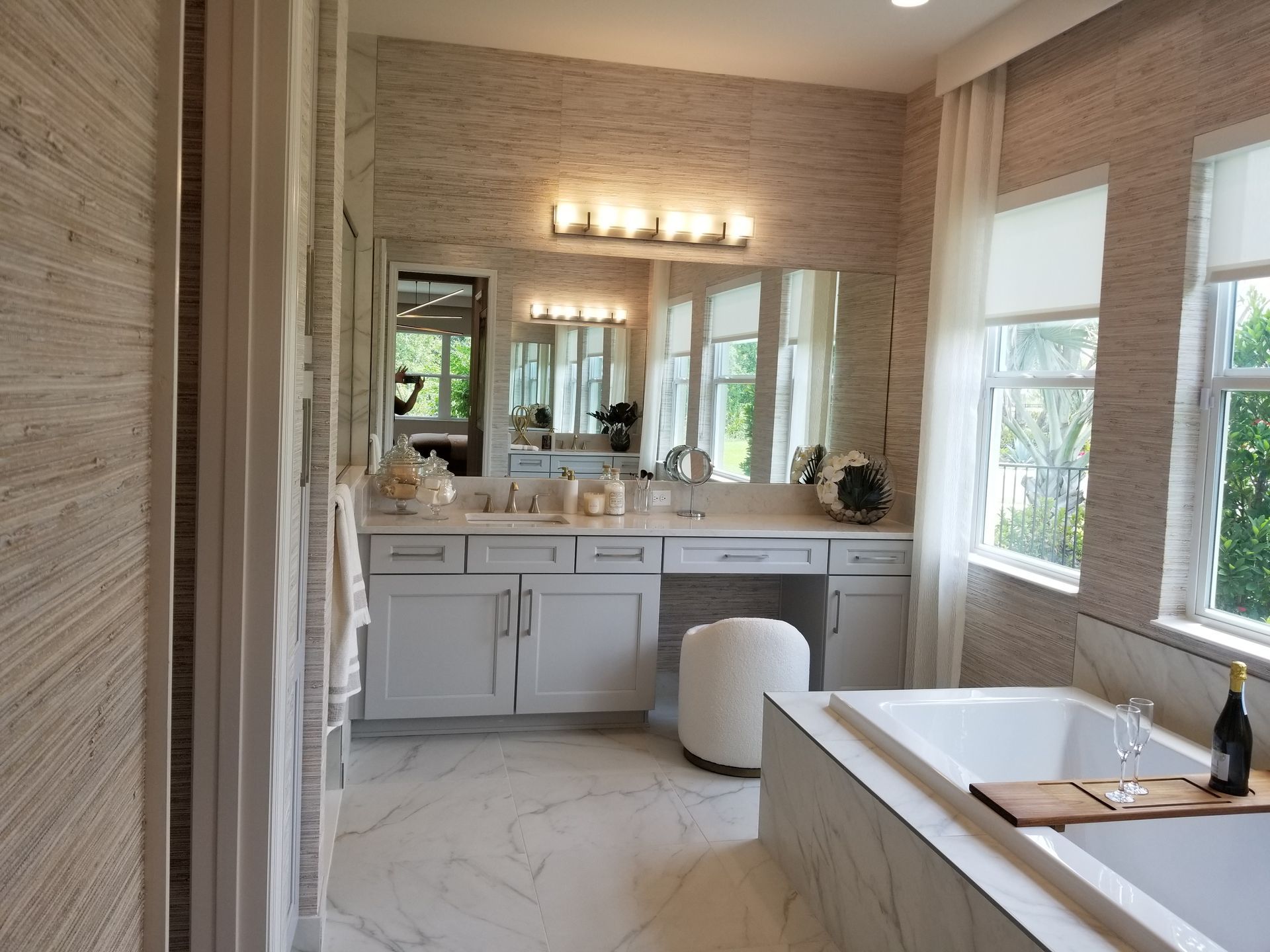Bright bathroom with marble walls, double vanity, mirror lights, and a freestanding tub by a window.
