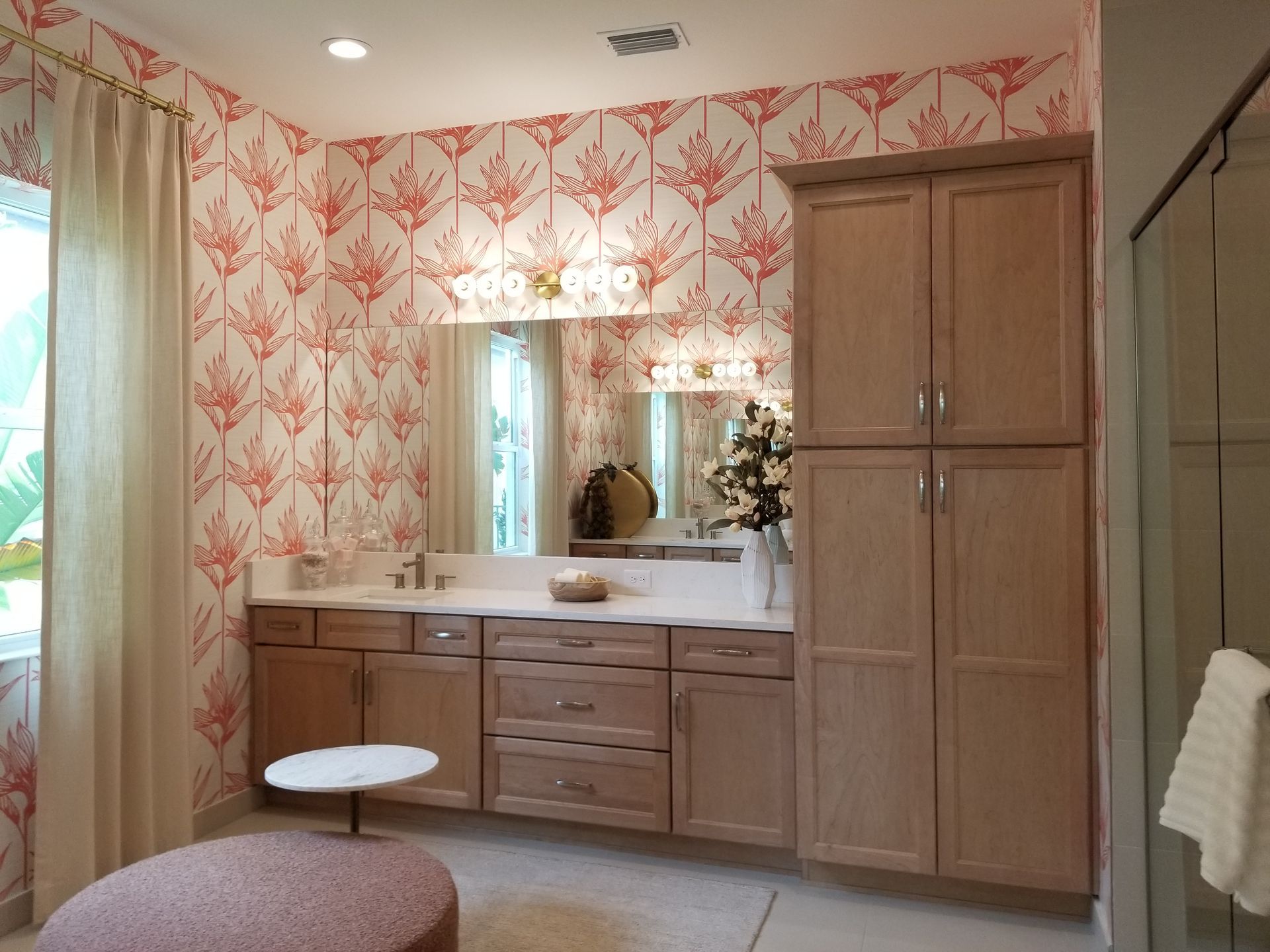 Bathroom vanity with wood cabinets, pink patterned wallpaper, mirror, and a small stool near the sink.