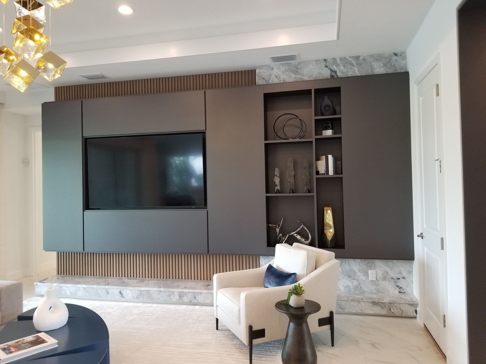 Modern living room with wall-mounted TV, dark wood shelving, and a white armchair by a window