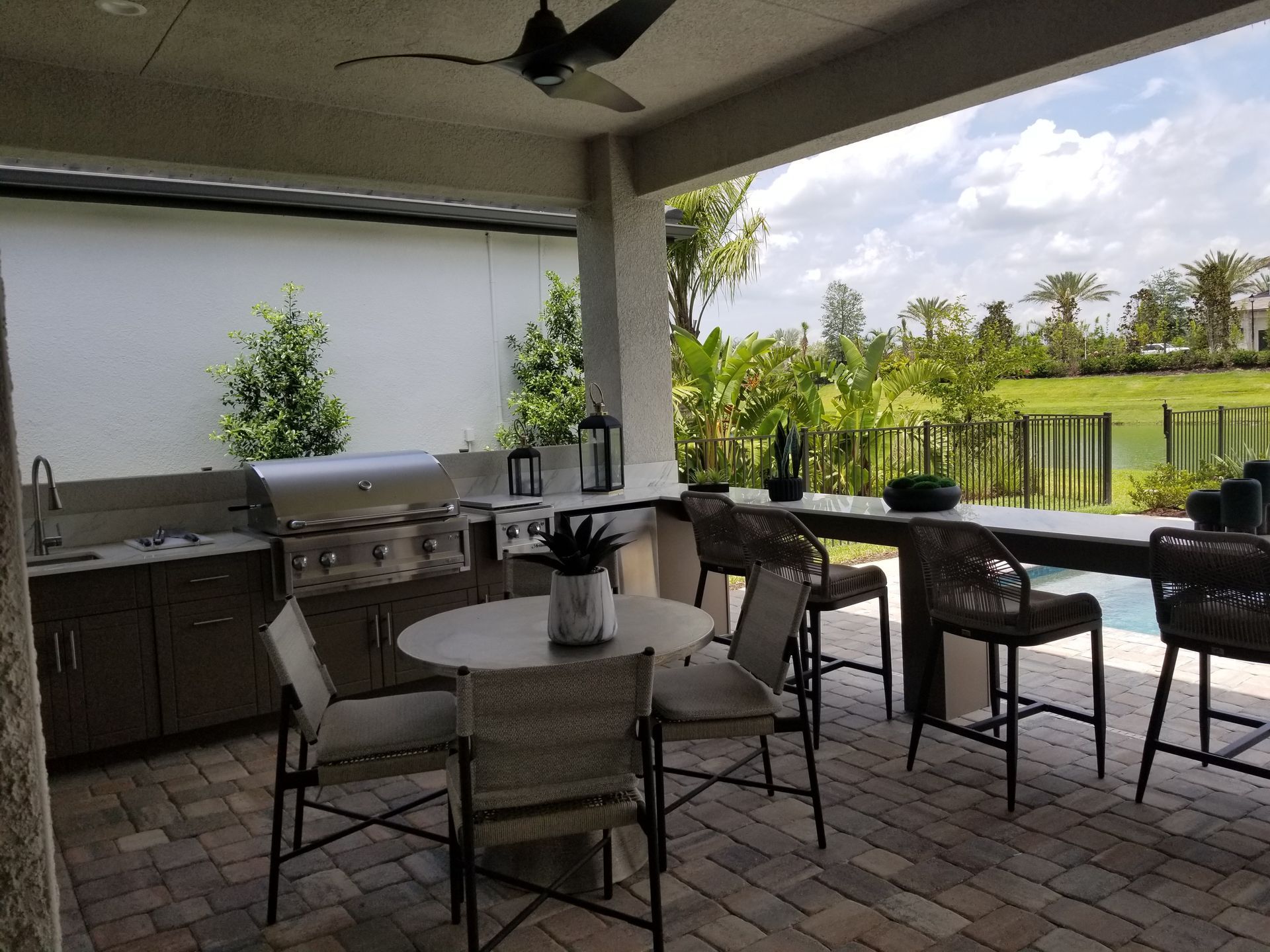 Covered patio with outdoor dining table, grill, bar stools, and a view of a pool and tropical landscaping.