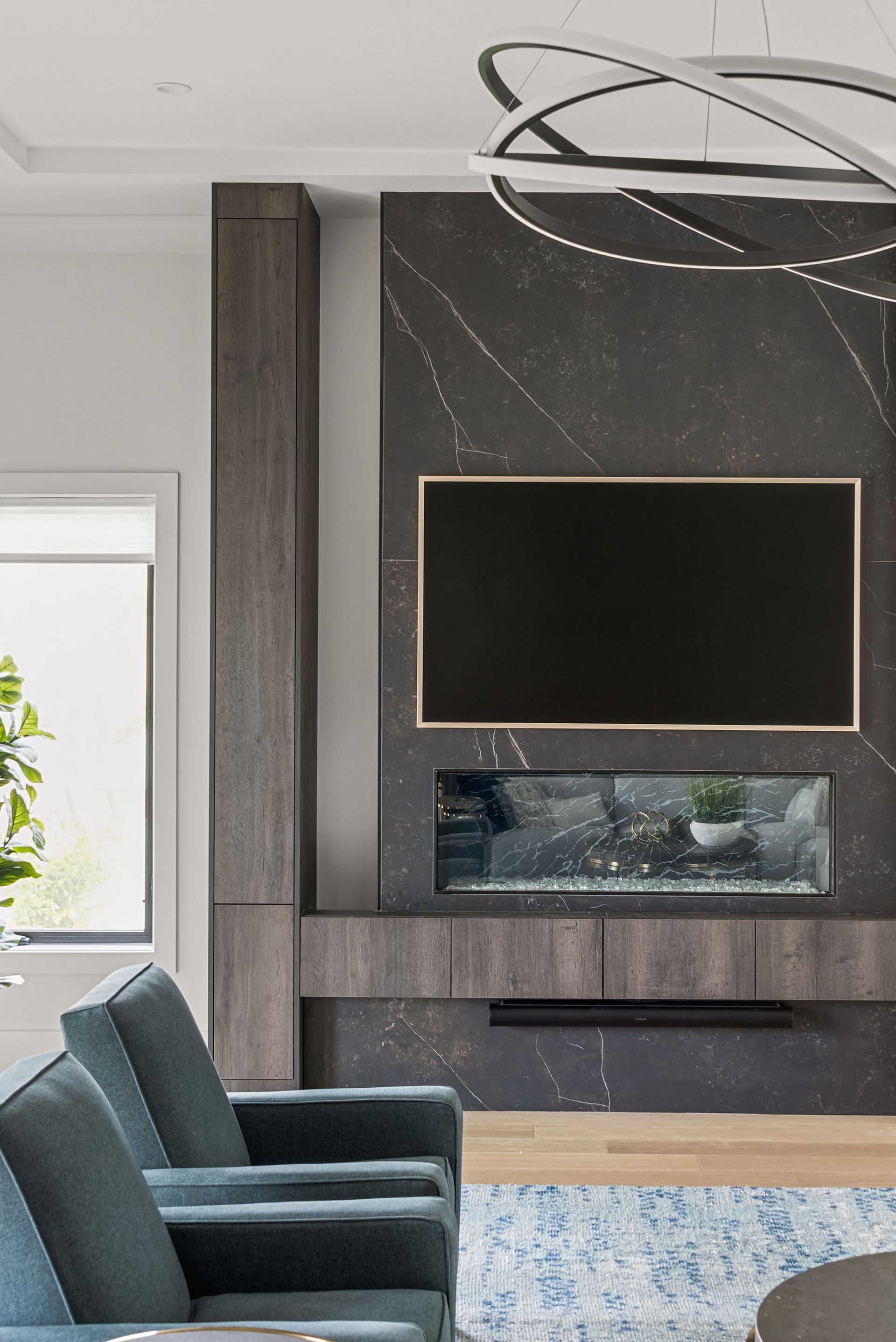 Modern living room with black fireplace wall, TV, teal chairs, and large window sunlight