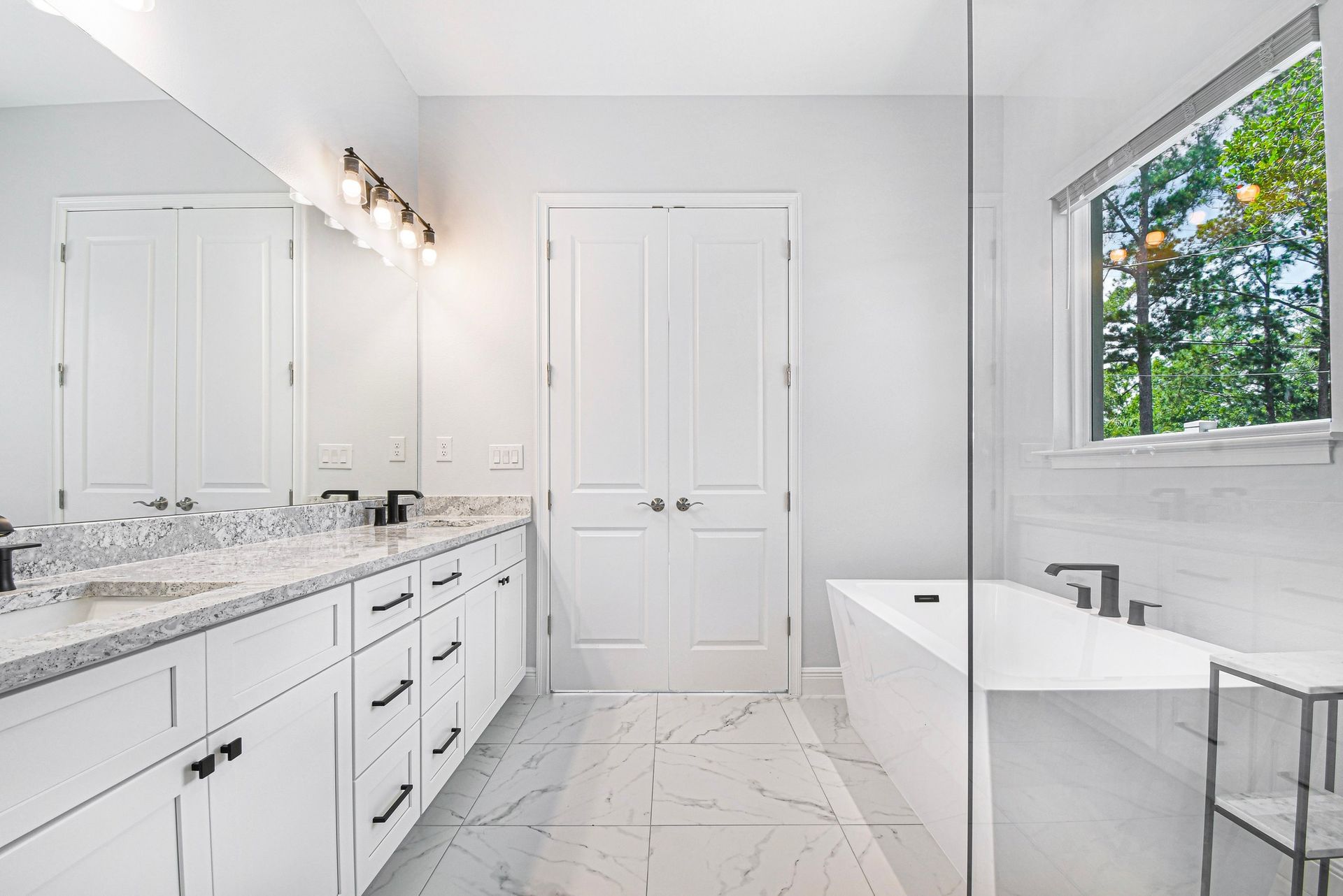 Bright white bathroom with double vanity, marble countertops, large mirror, and window view of greenery