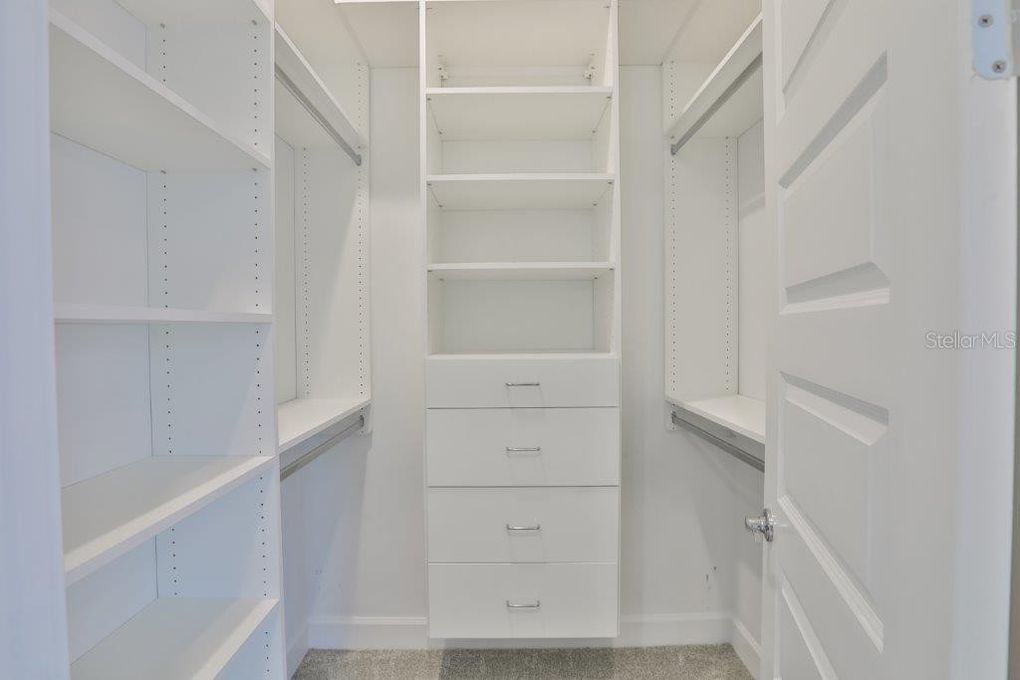 Narrow white walk-in closet with shelves, drawers, hanging rods, and a white door.