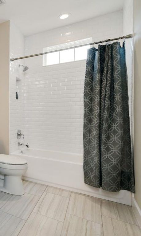 White bathroom with tub-shower, subway tile, and dark patterned curtain; toilet at left.
