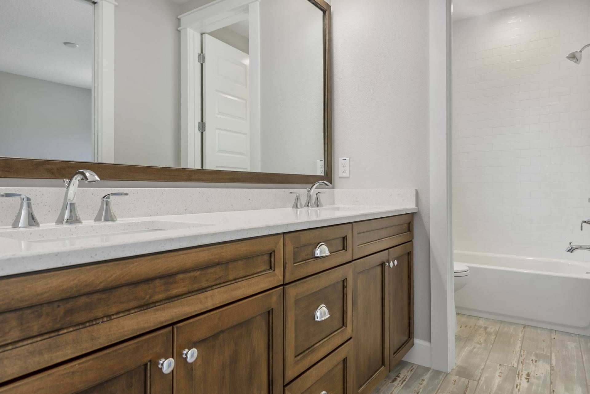 Bathroom vanity with white countertop, wooden cabinets, mirror, and tub in the background