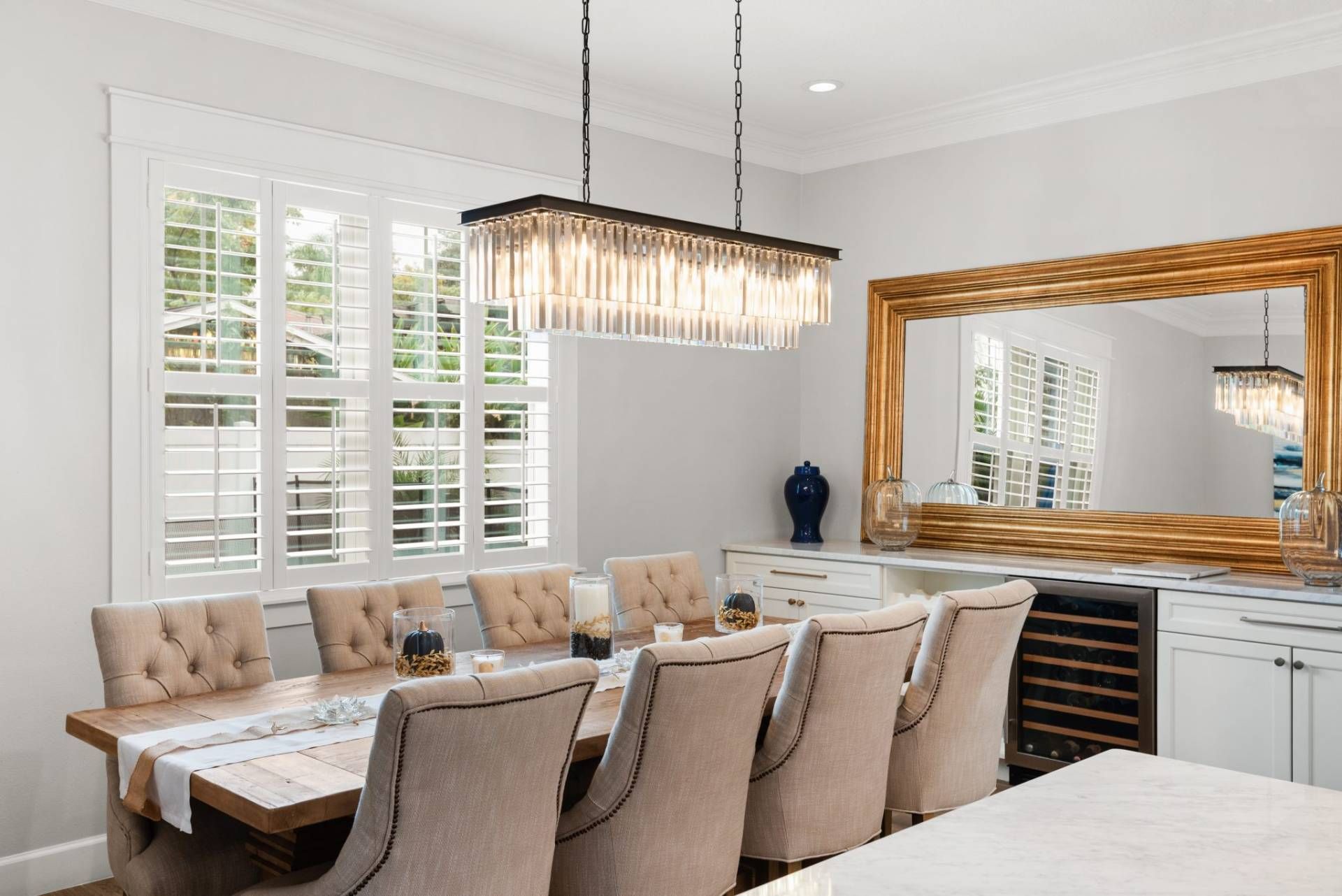 Bright dining room with a large table, upholstered chairs, chandelier, and gold-framed mirror.