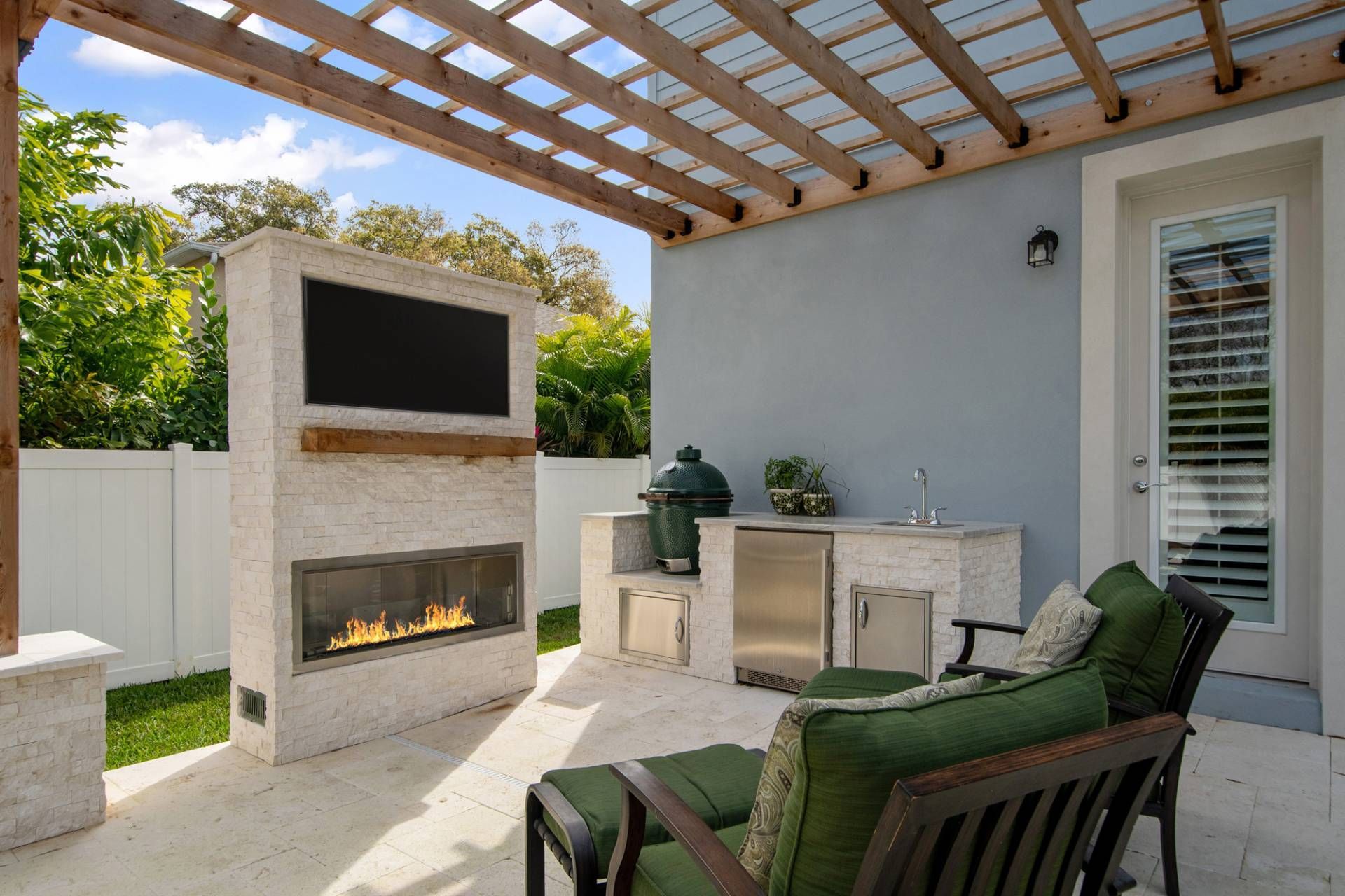 Covered patio with a built-in fireplace, outdoor TV, and cushioned seating next to a white fence