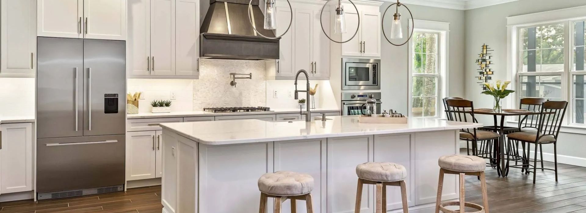 Bright modern kitchen with white island, stainless steel fridge, pendant lights, and dining area by windows